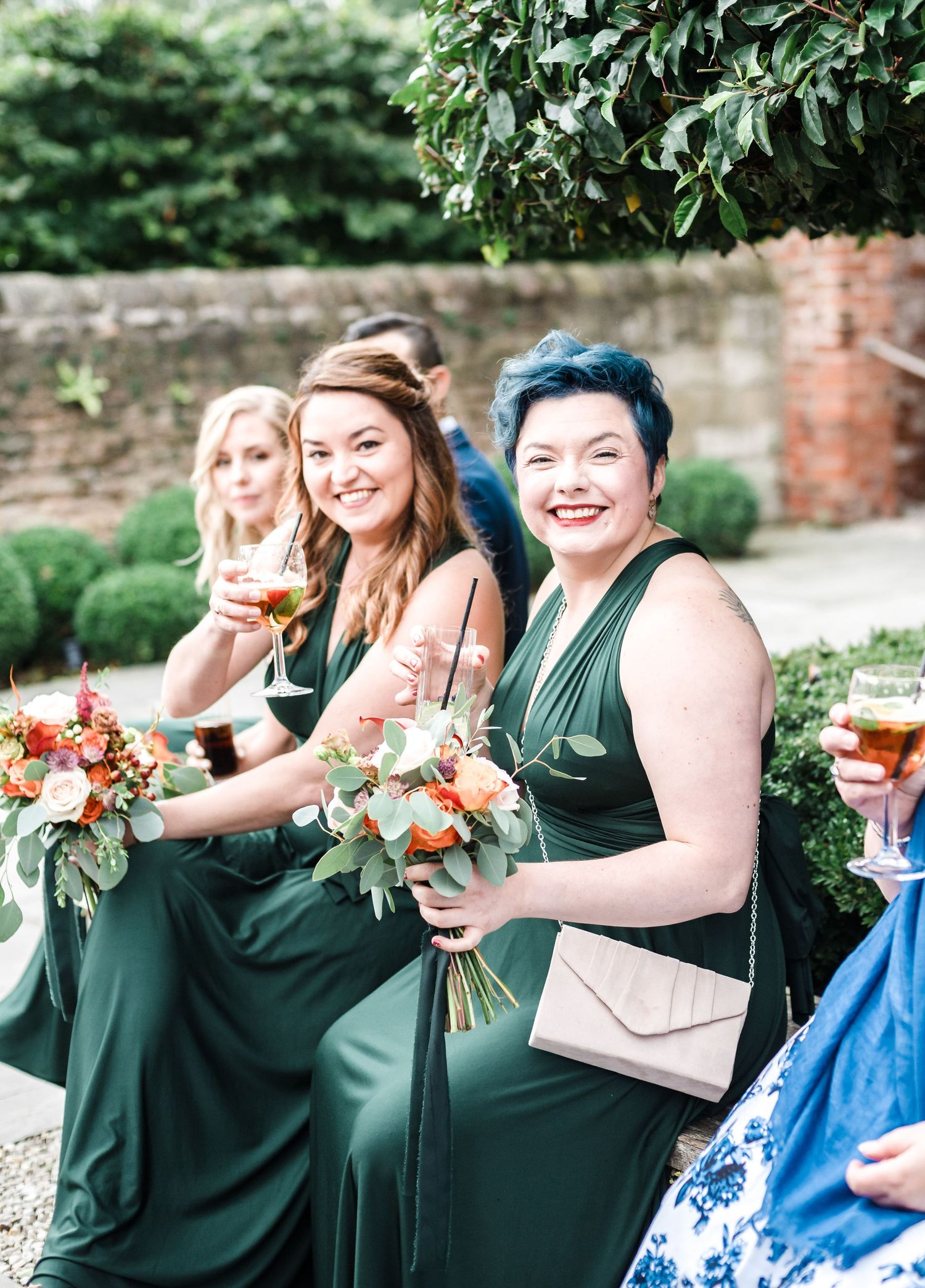 Wedding guests in green dresses hold bouquets and champagne at outdoor reception.