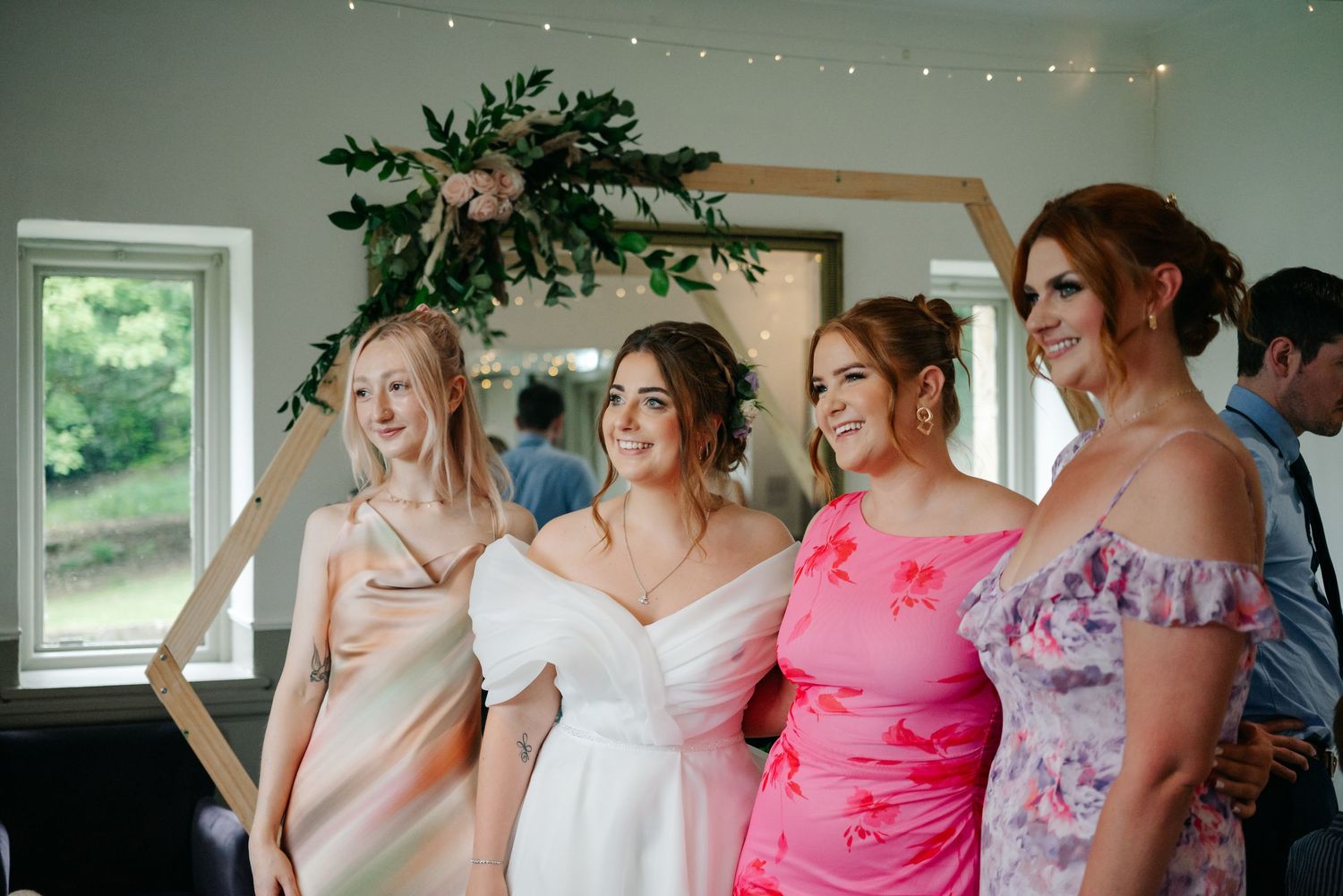 A group of women in elegant dresses smile and laugh together at a formal event by a window.