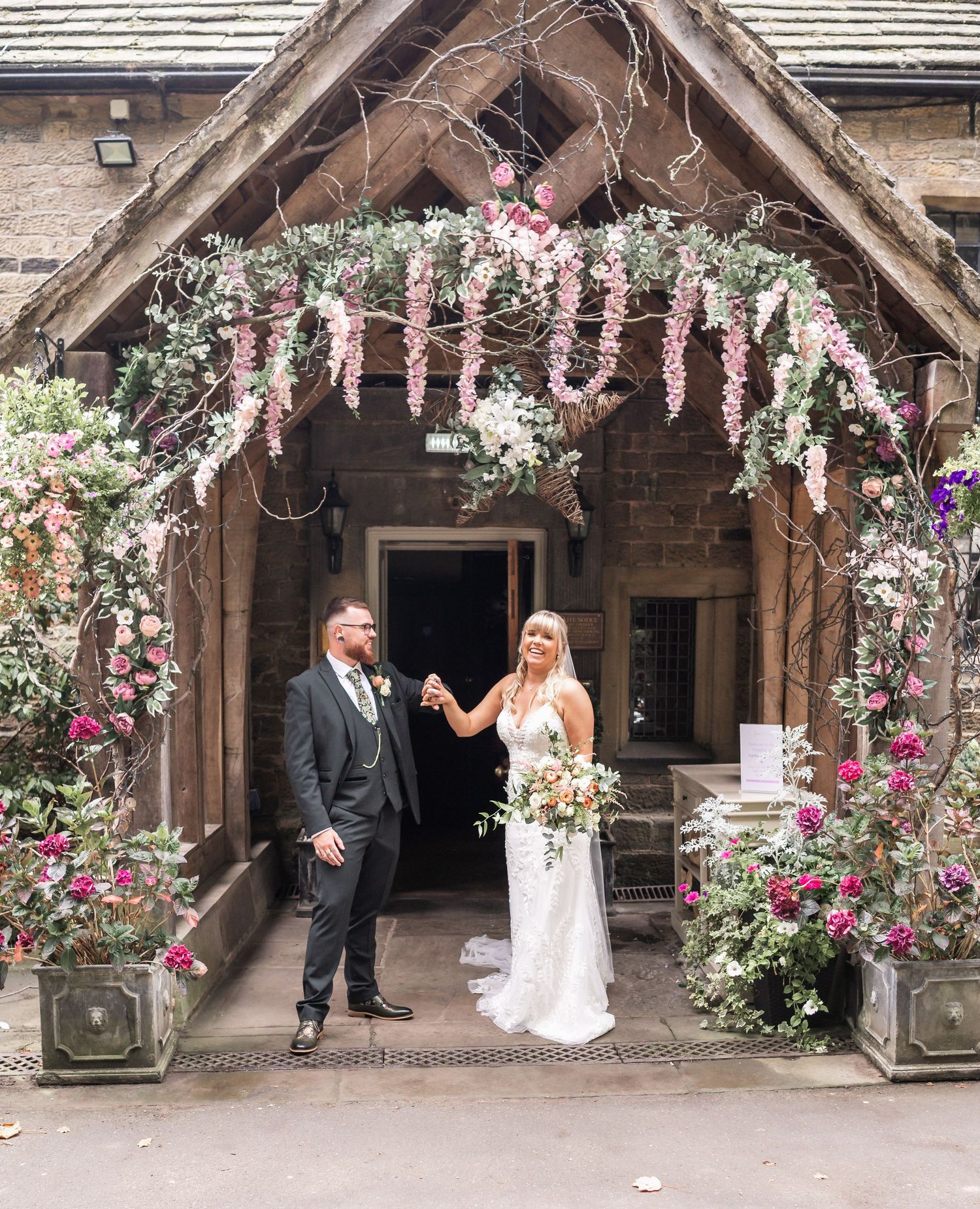 Couple standing under floral archway decorated with pink blooms and greenery.