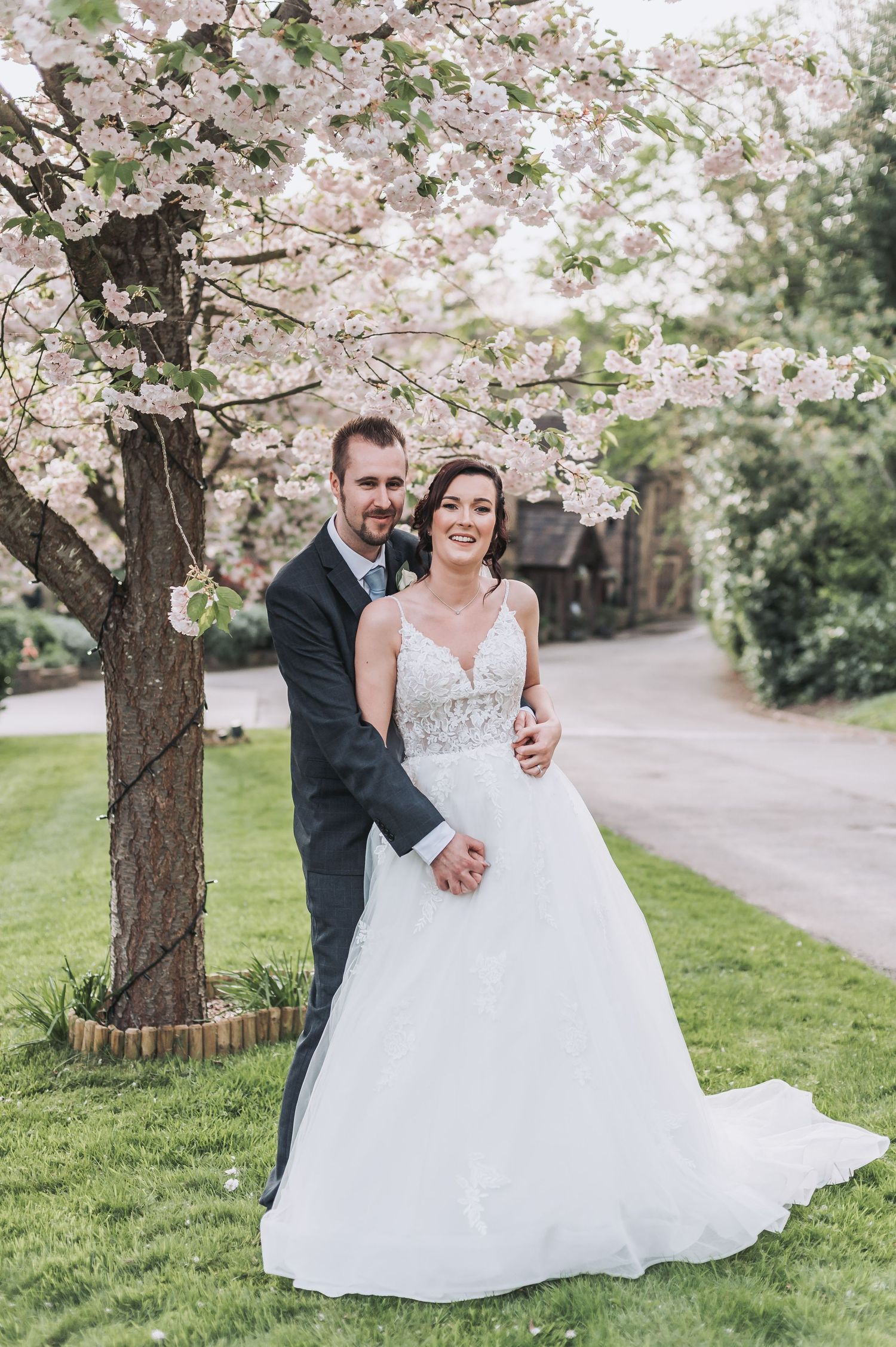 Wedding couple walking along a path under spring cherry blossoms in formal attire.