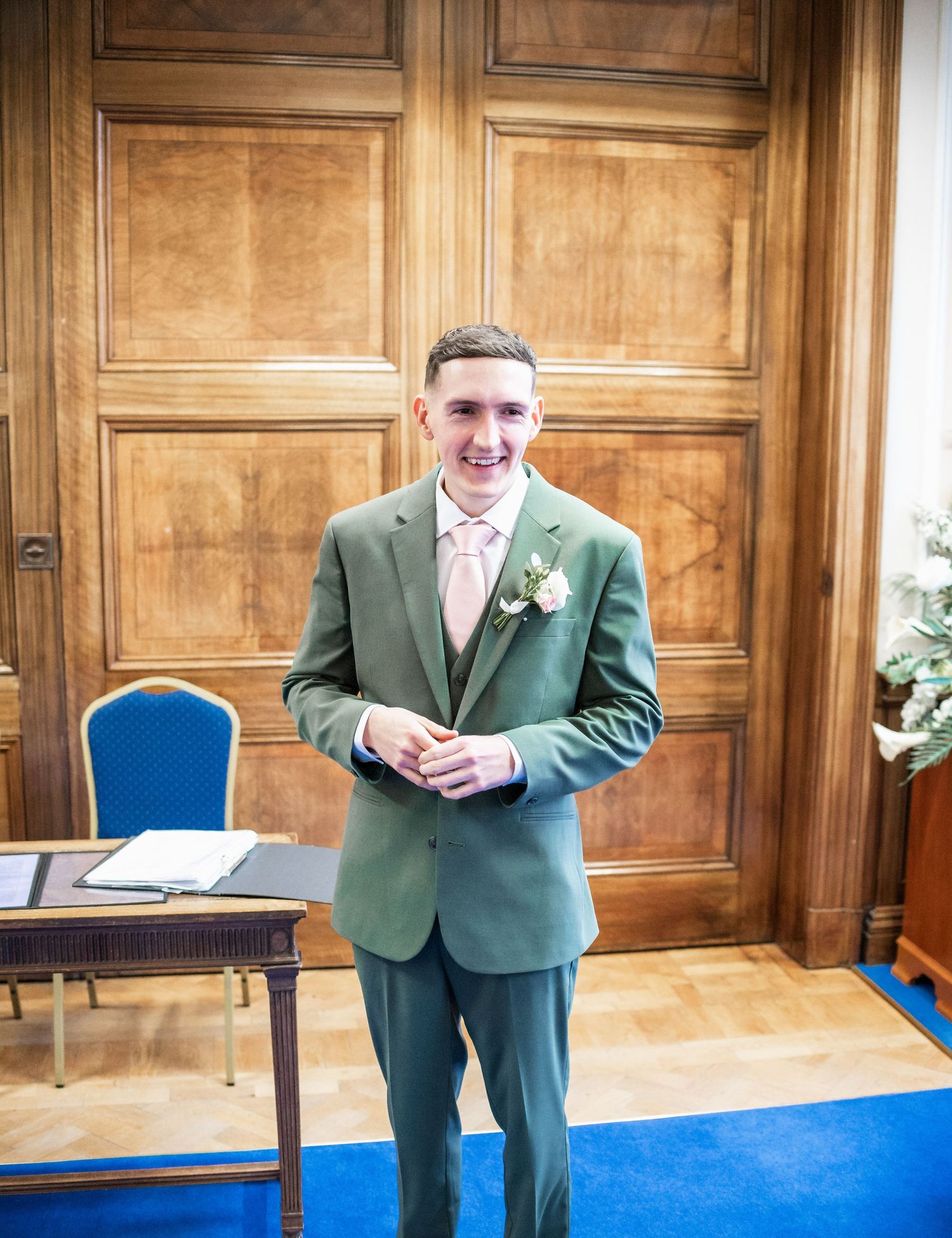 Groom in mint green suit with pink tie stands in front of wooden paneled doors.
