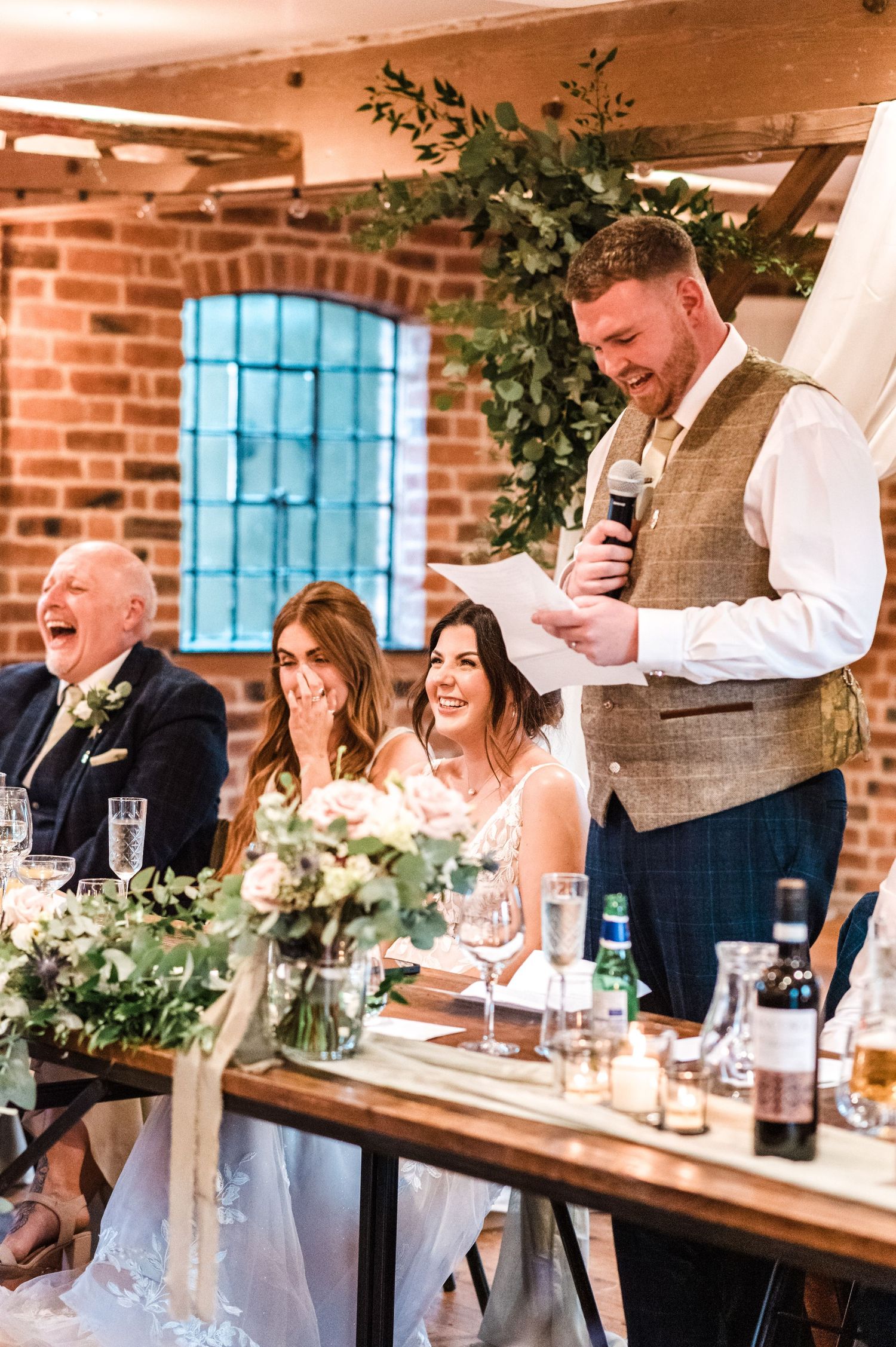 Wedding speech being delivered at head table with guests laughing in rustic venue.