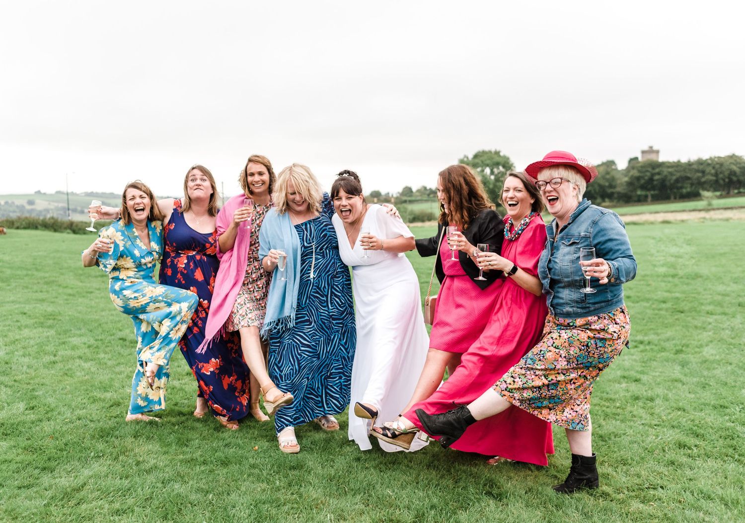 Group of wedding guests in colorful dresses posing playfully on grass.