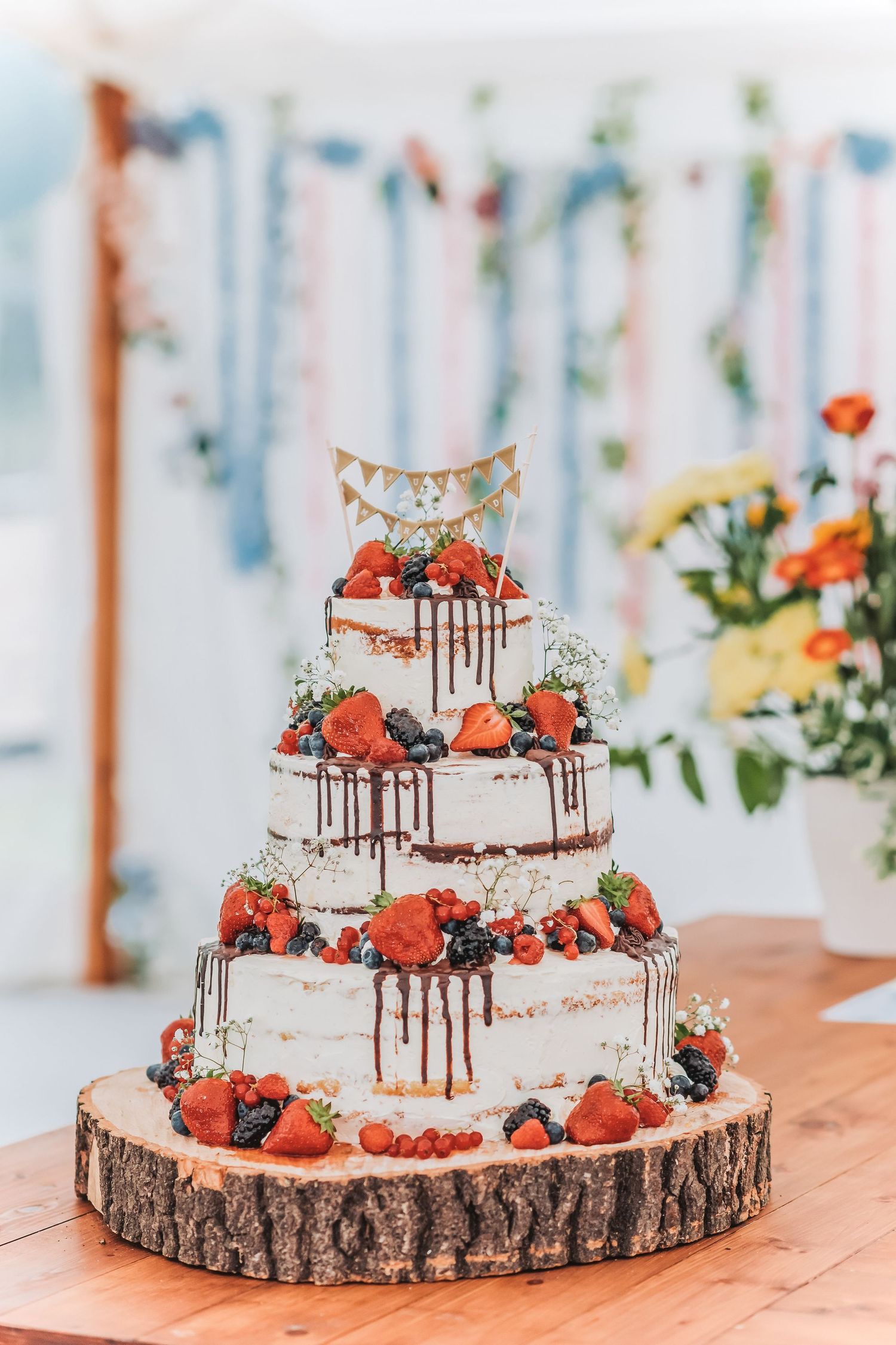 Rustic naked wedding cake decorated with fresh strawberries and flowers on wood slice base.