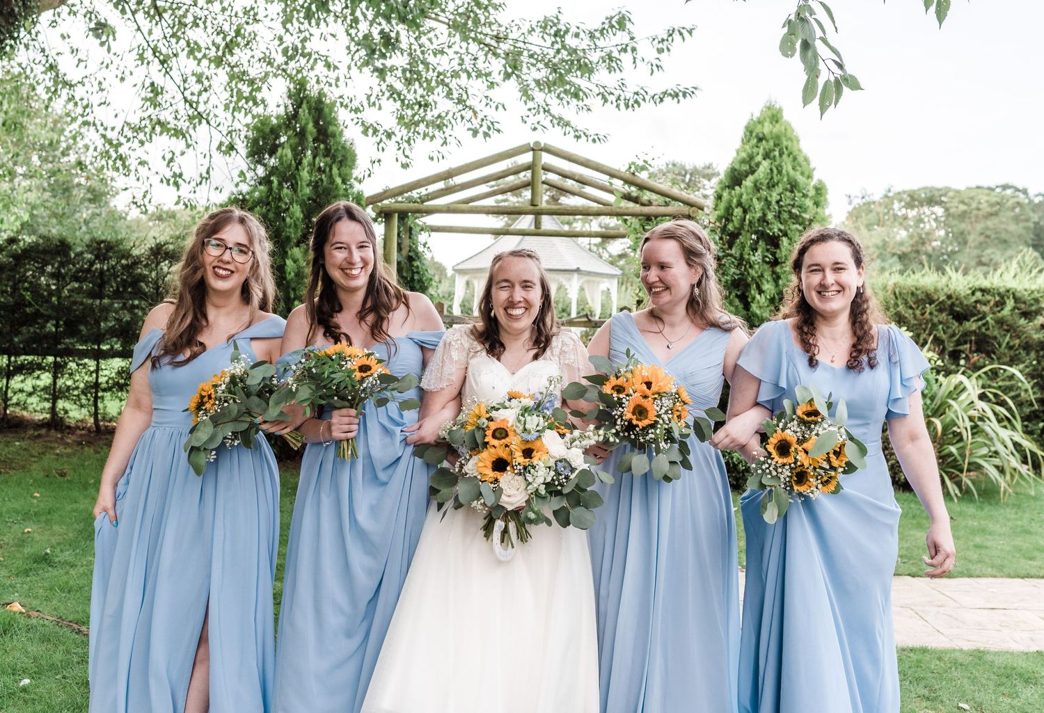 Bridal party in light blue dresses holding sunflower bouquets pose outdoors under green trees.