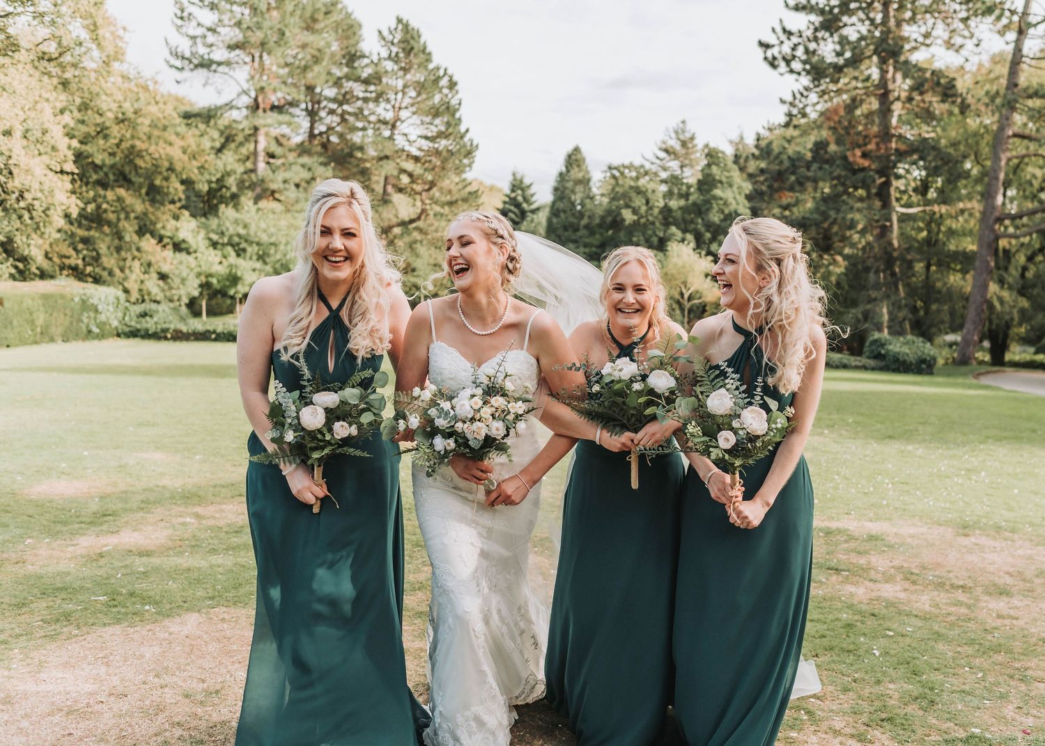 Wedding party wearing elegant emerald green dresses pose with bouquets in an outdoor garden setting.