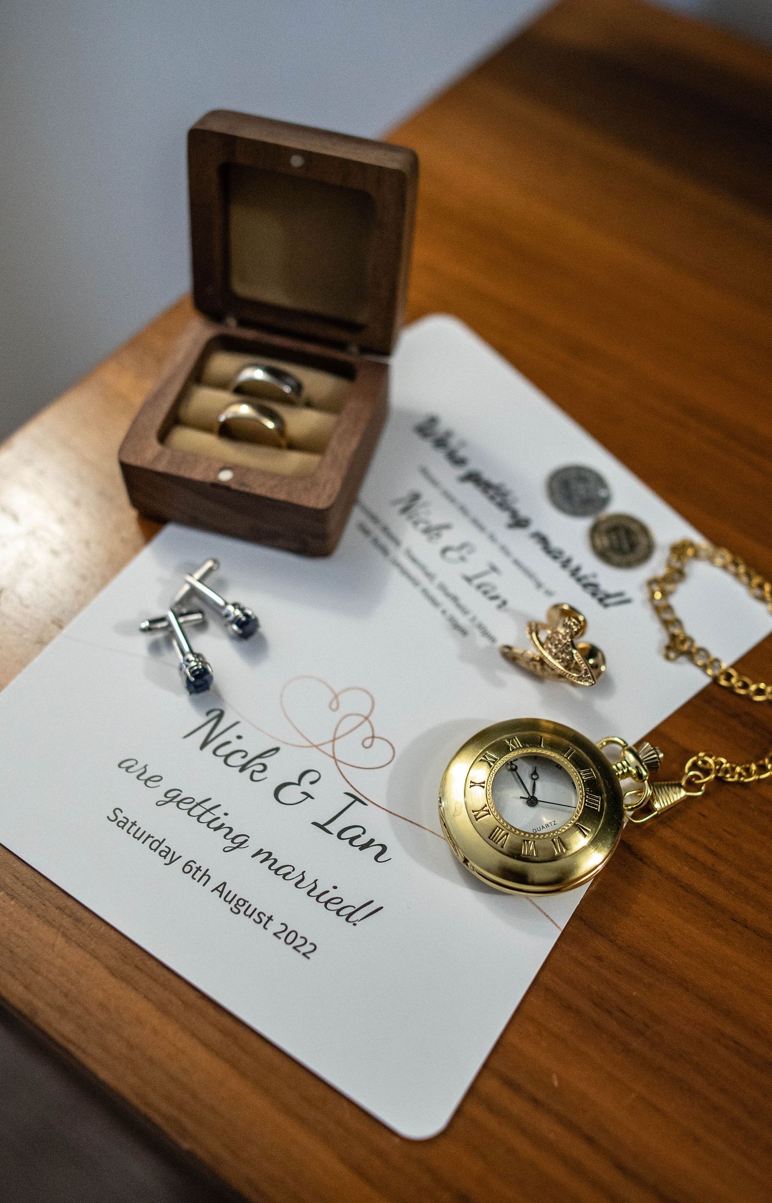 Wedding rings in wooden box with vintage pocket watch and invitation details.