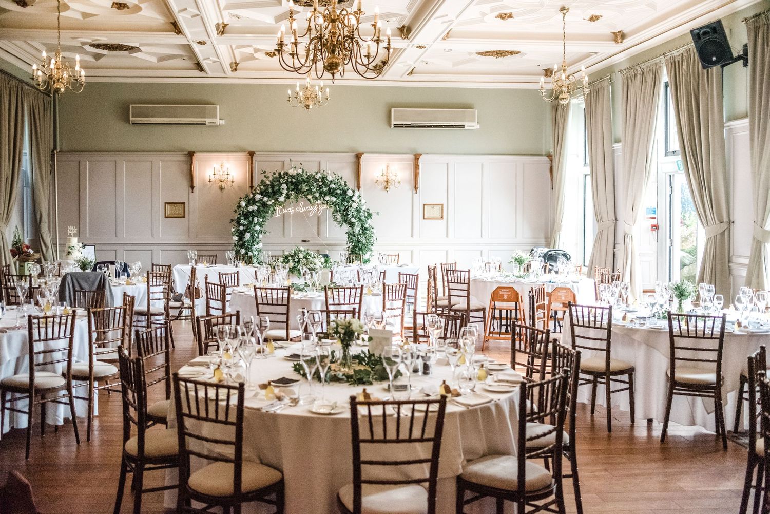 Elegant wedding reception venue with chandeliers and round tables decorated in white and greenery.