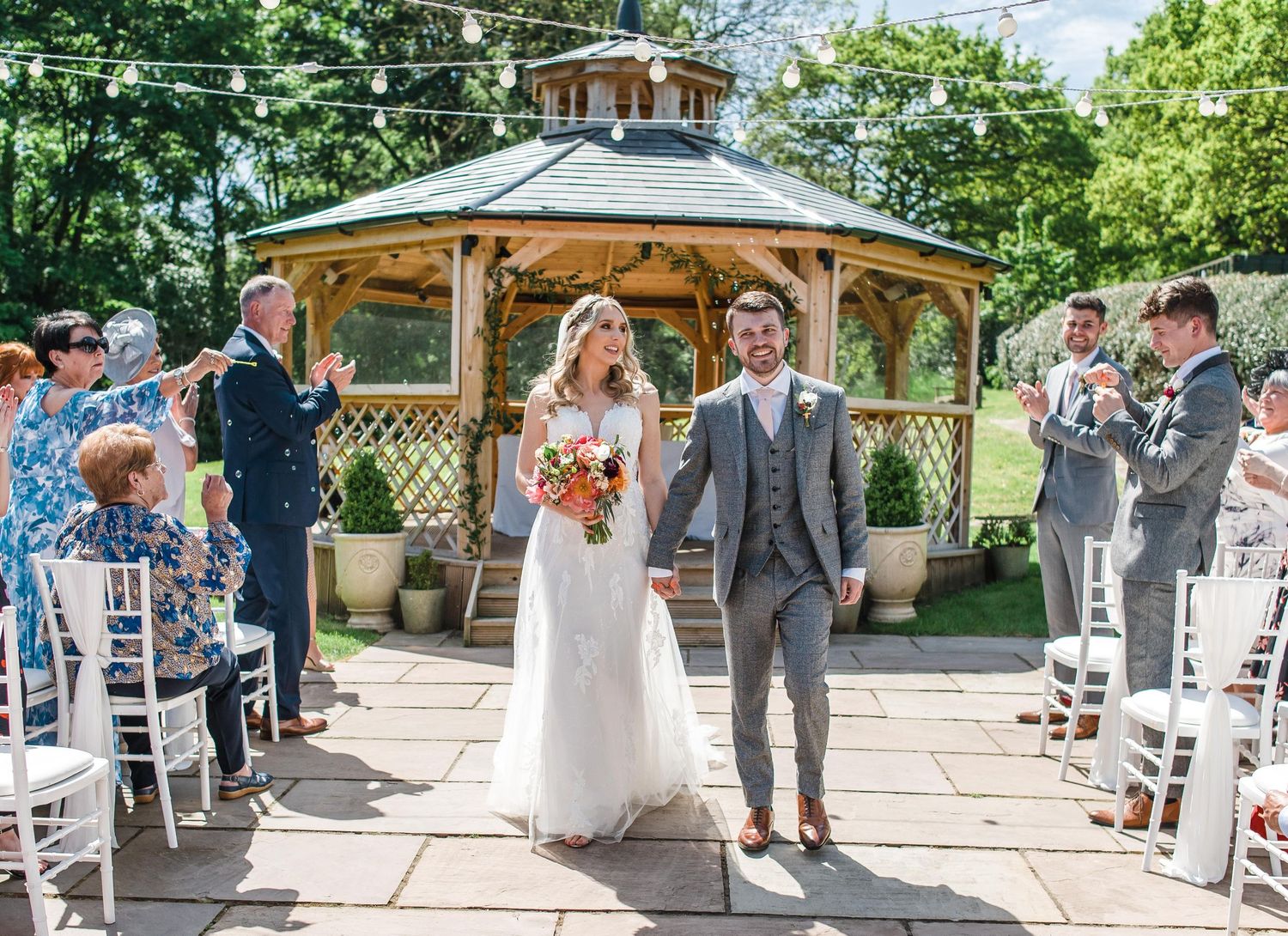 Outdoor wedding ceremony at a gazebo with guests seated in white chairs on a stone patio.