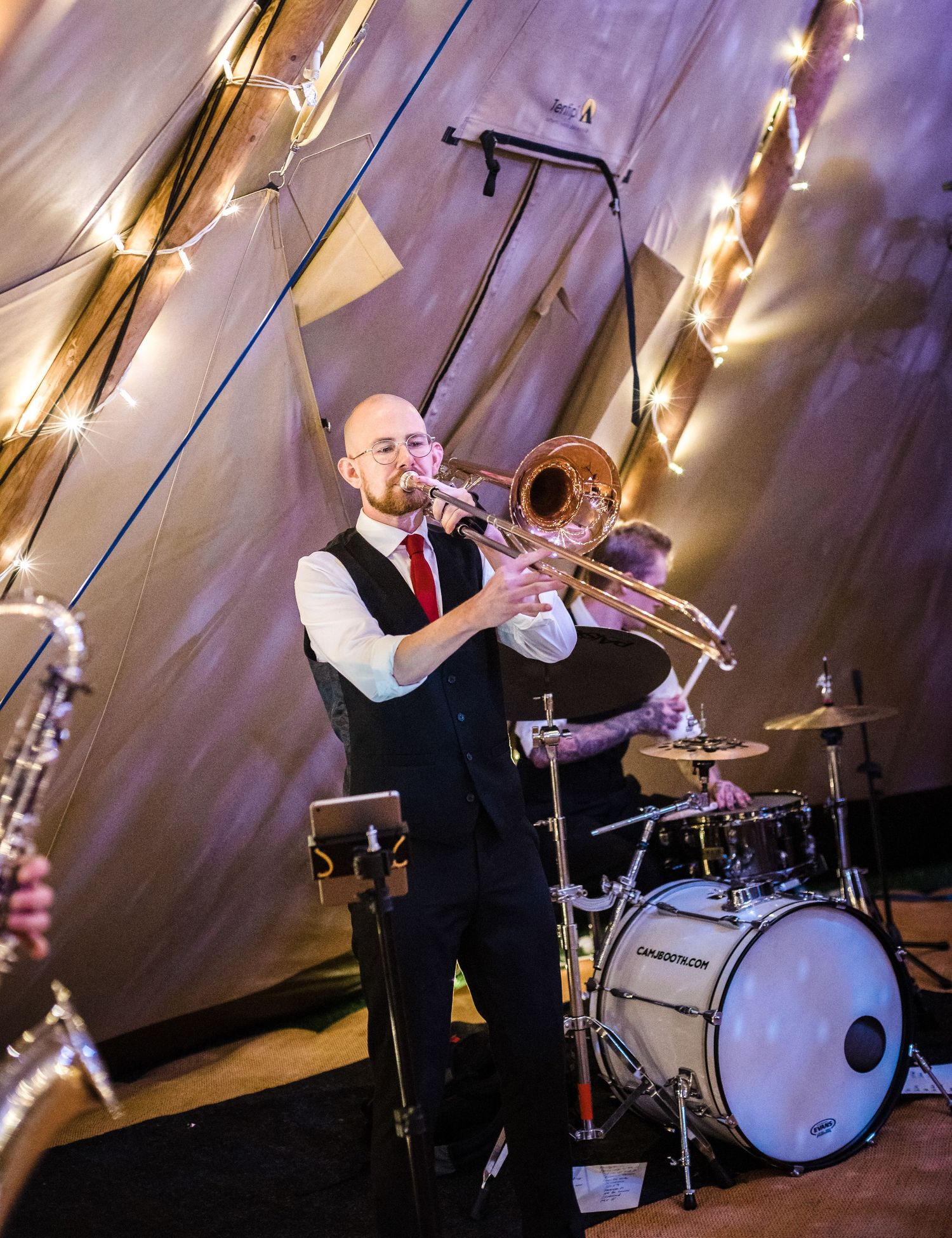 Professional musician playing trombone during a wedding reception in a tent with string lights.