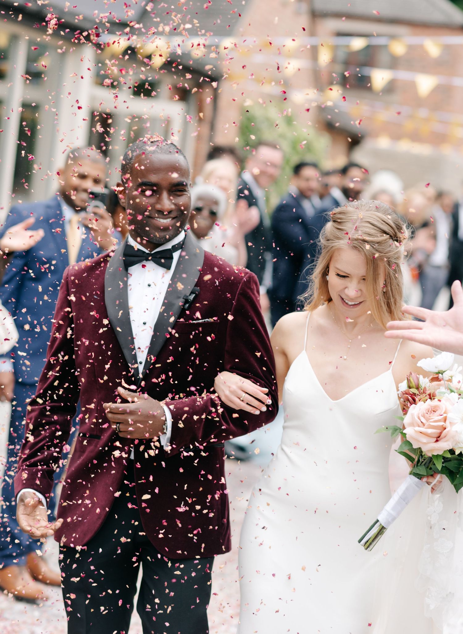 Bride and groom walk through colorful confetti shower, groom in burgundy velvet tuxedo, bride in white gown holding pink bouquet.