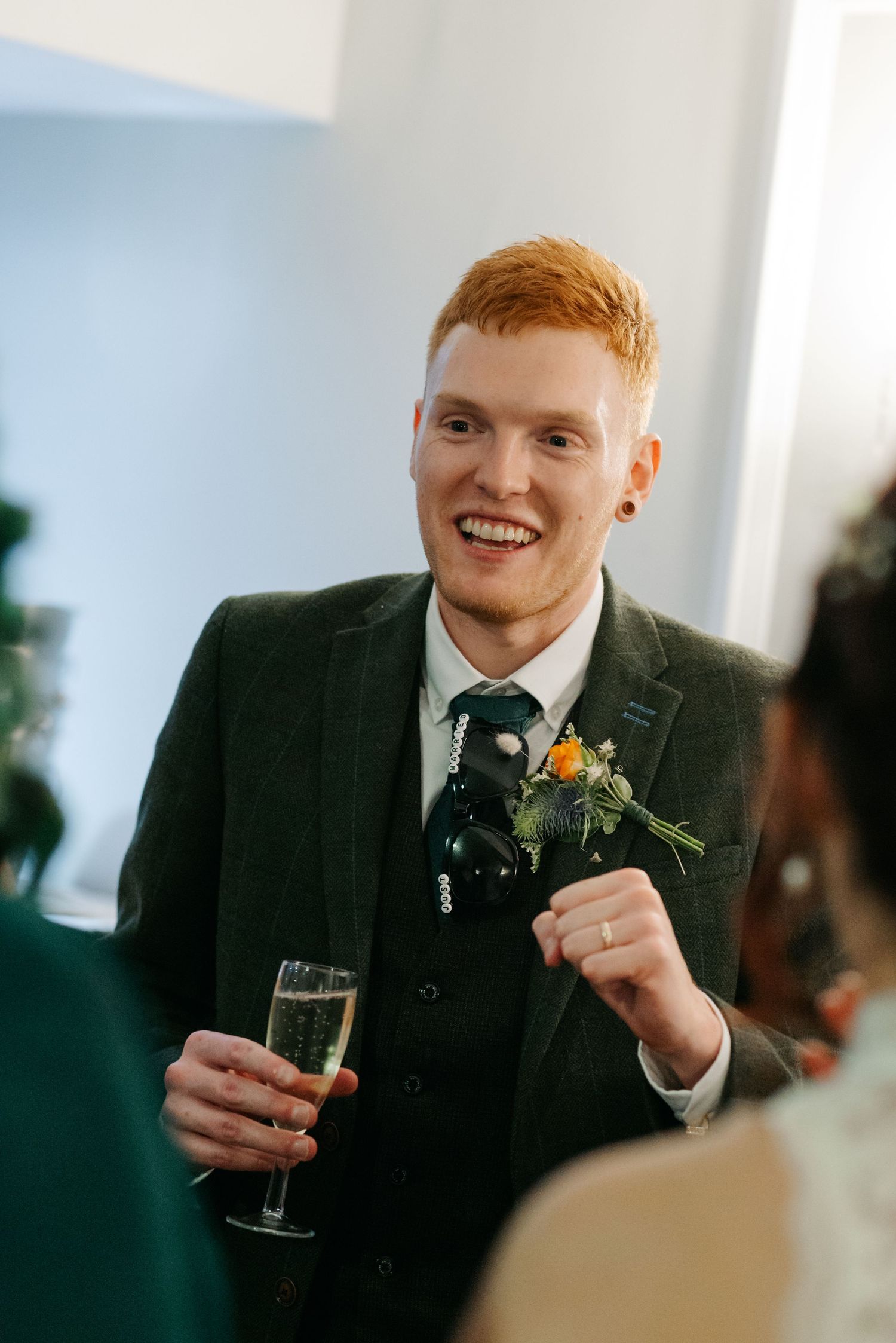 Person in dark suit with boutonniere smiling while holding champagne glass.