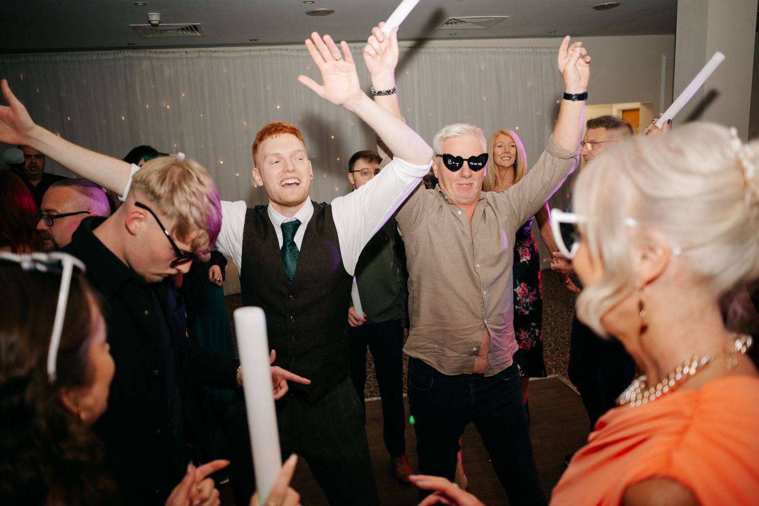Energetic wedding reception dance party with guests raising their hands in celebration.