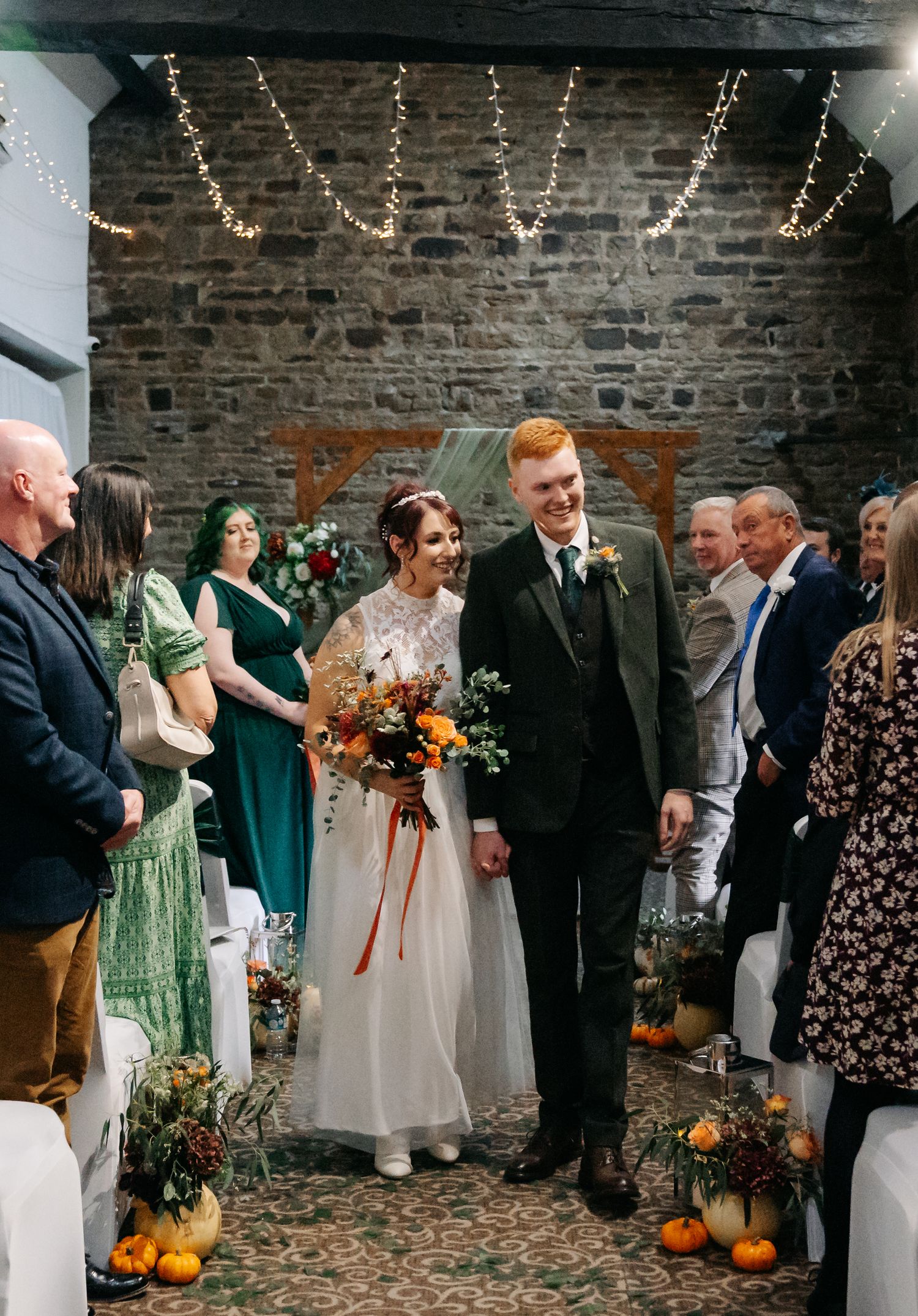 Wedding guests gather outside a brick building decorated with pumpkins and fall decor.