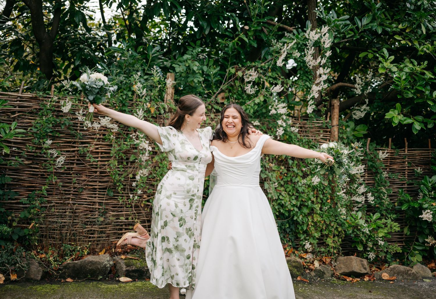 Two people in white dresses celebrate joyfully in a garden setting with a woven fence.