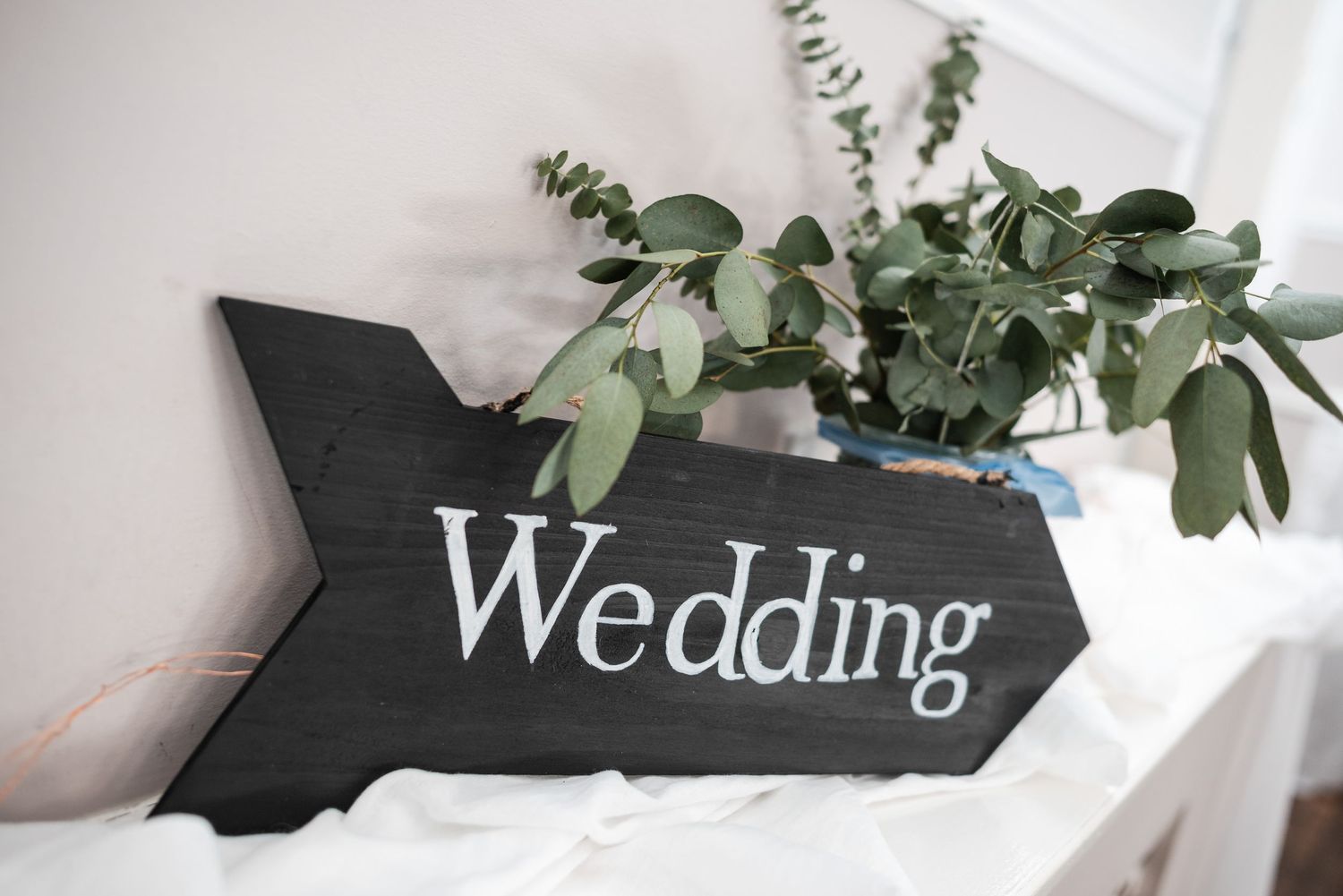 A black wooden arrow sign with white 'Wedding' text sits beside green eucalyptus leaves against a white background.