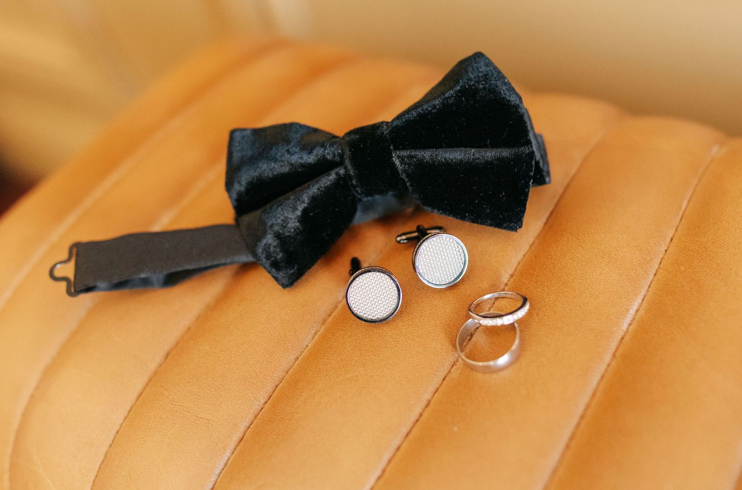 Black velvet bow tie, silver cufflinks, and wedding rings arranged on tan leather upholstery.