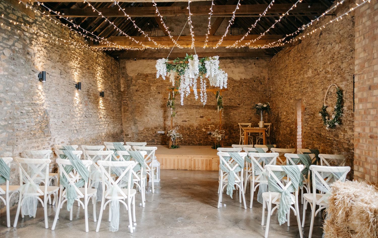 Rustic barn wedding venue interior decorated with string lights and white chairs for ceremony setup.