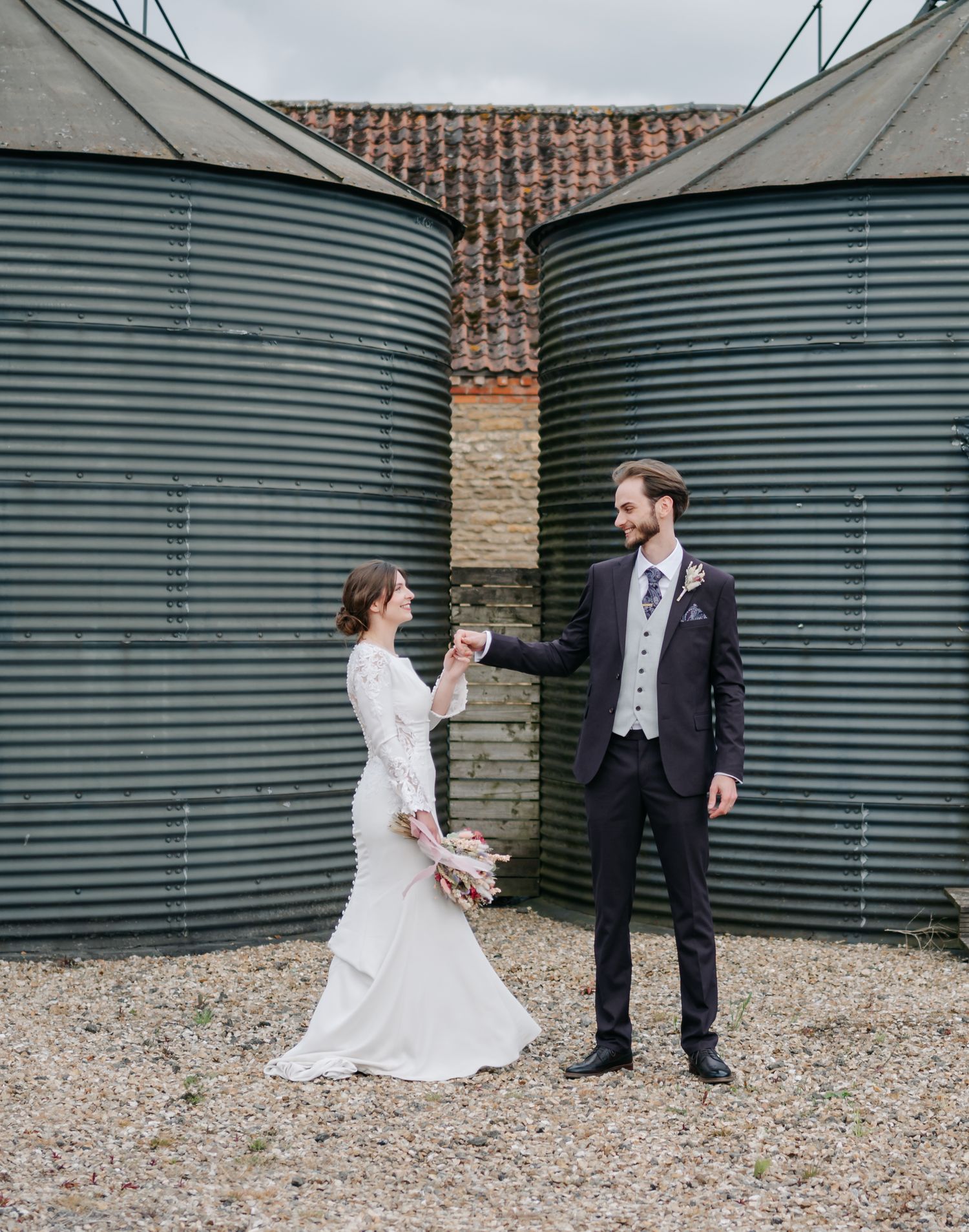 Couple shares playful moment between industrial storage tanks at wedding venue.