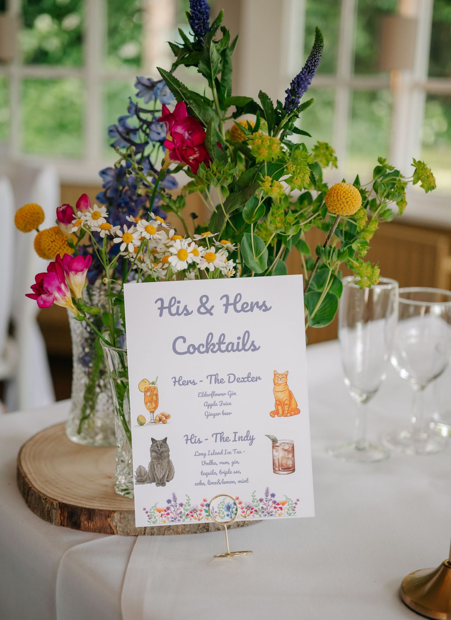 His and Hers cocktail menu with cat illustrations displayed on rustic wooden centerpiece with wildflowers.