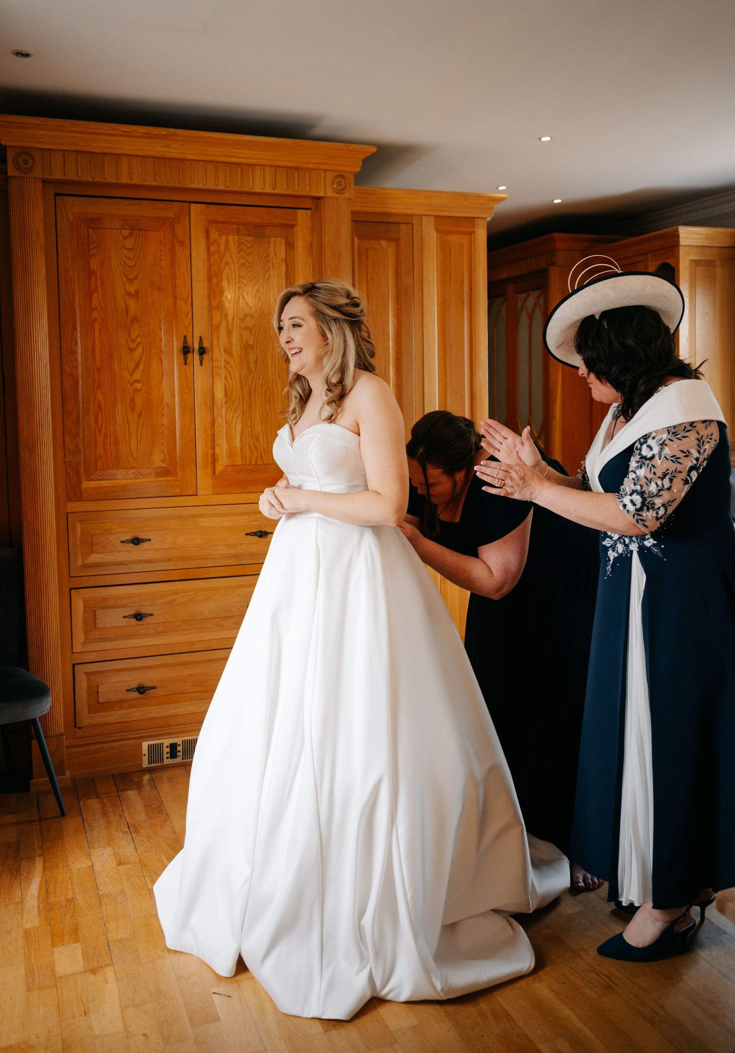 A bride in a classic white wedding gown getting final adjustments in a wood-paneled dressing room.
