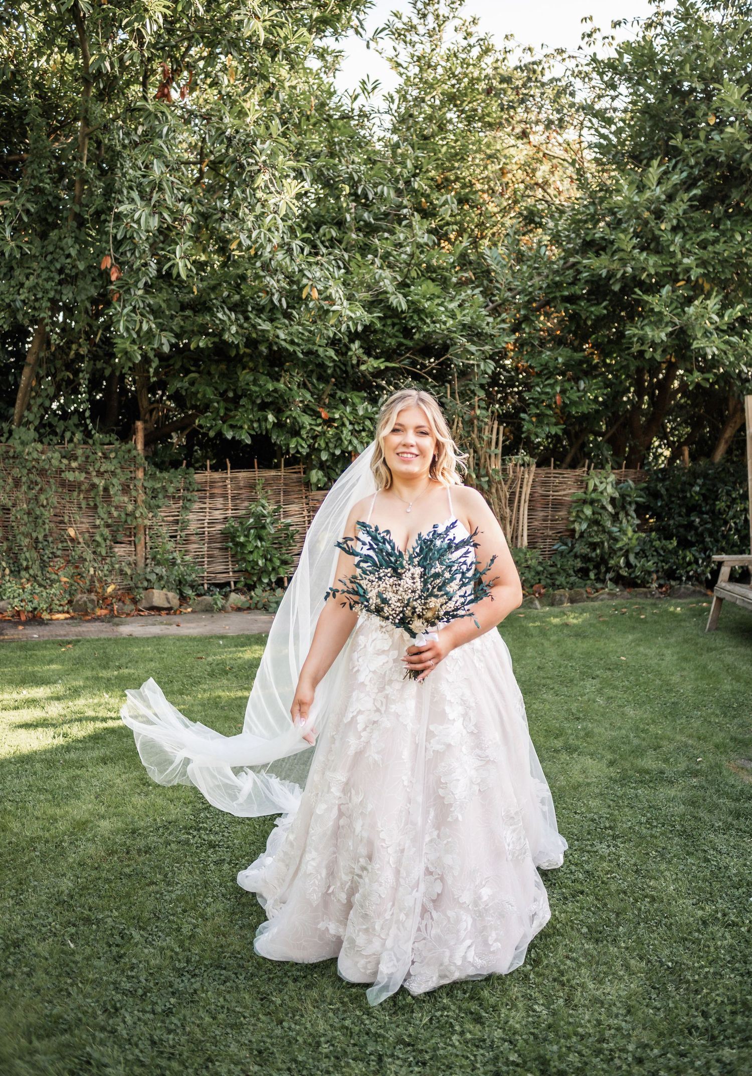 A flowing white wedding dress catches the breeze in a garden setting with green foliage.