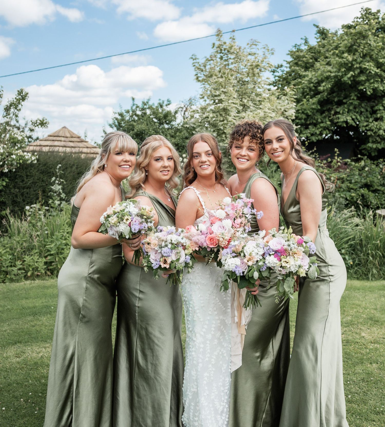 Bridal party poses together outdoors holding colorful summer bouquets against garden backdrop.