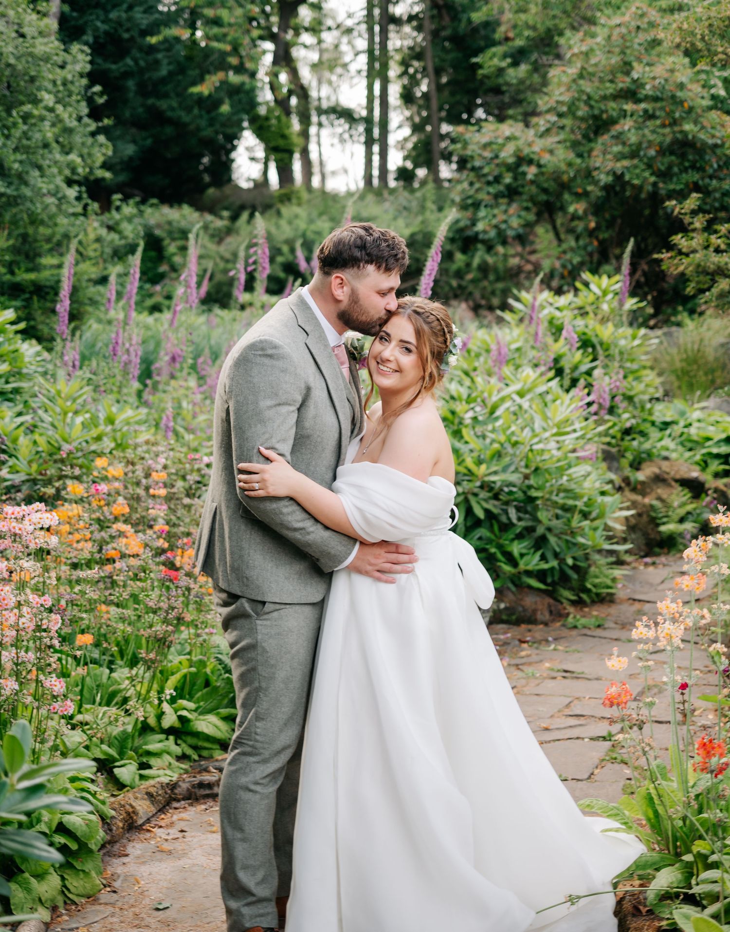 A newlywed couple shares an intimate moment in a colorful garden with purple and orange flowers.