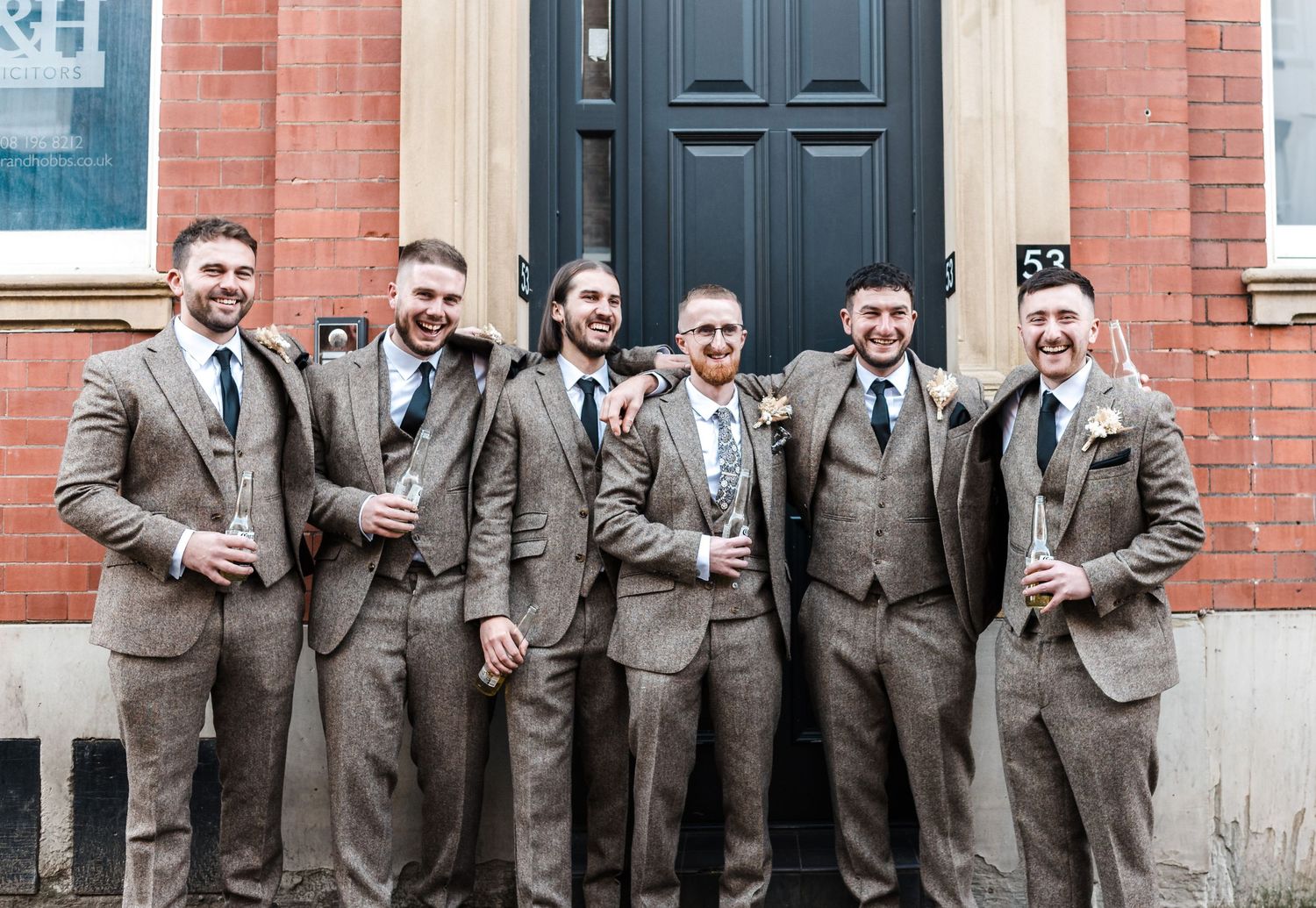 Groomsmen in matching tweed suits pose against a red brick wall.