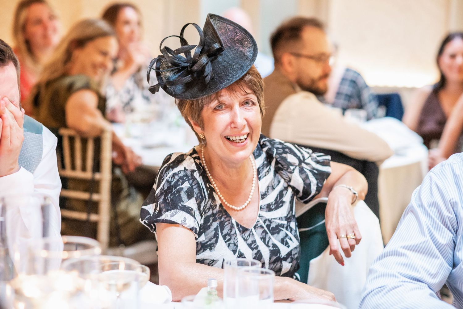 Wedding guest in navy fascinator hat smiling at reception table.