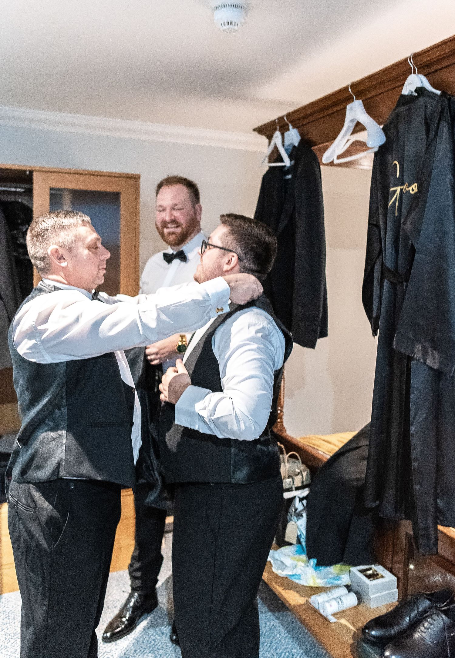 Groomsmen helping each other get dressed in formal attire before a wedding ceremony.