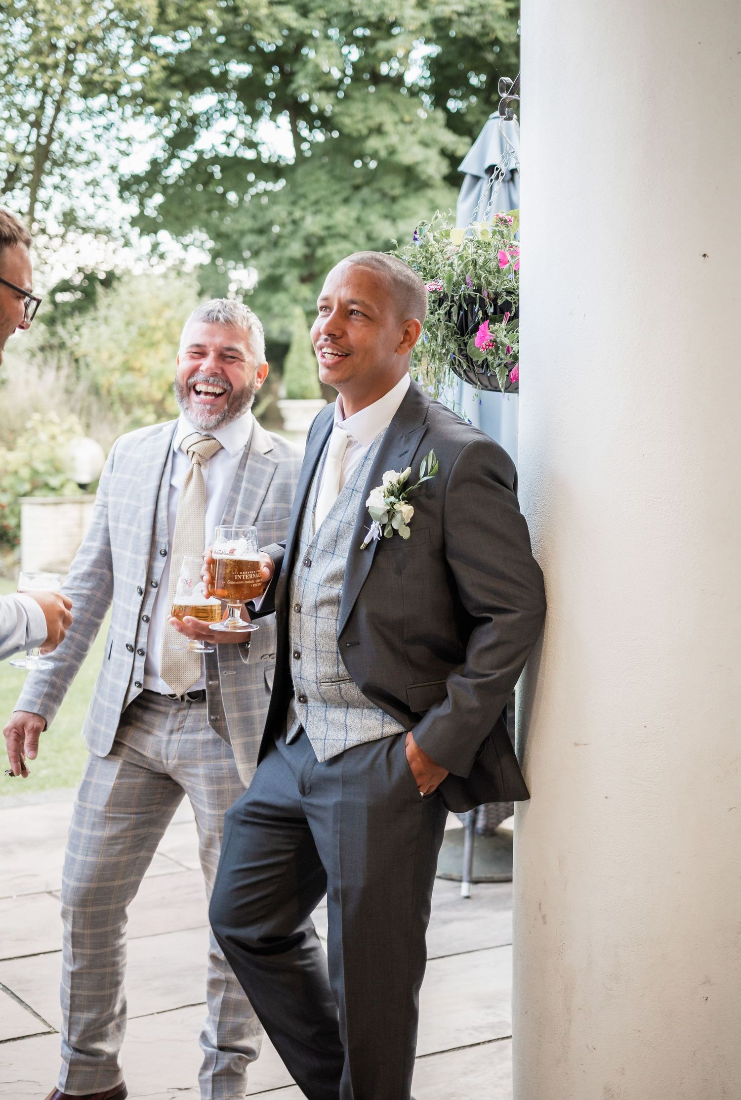 Two wedding guests in gray suits share a laugh while enjoying drinks on an outdoor patio.