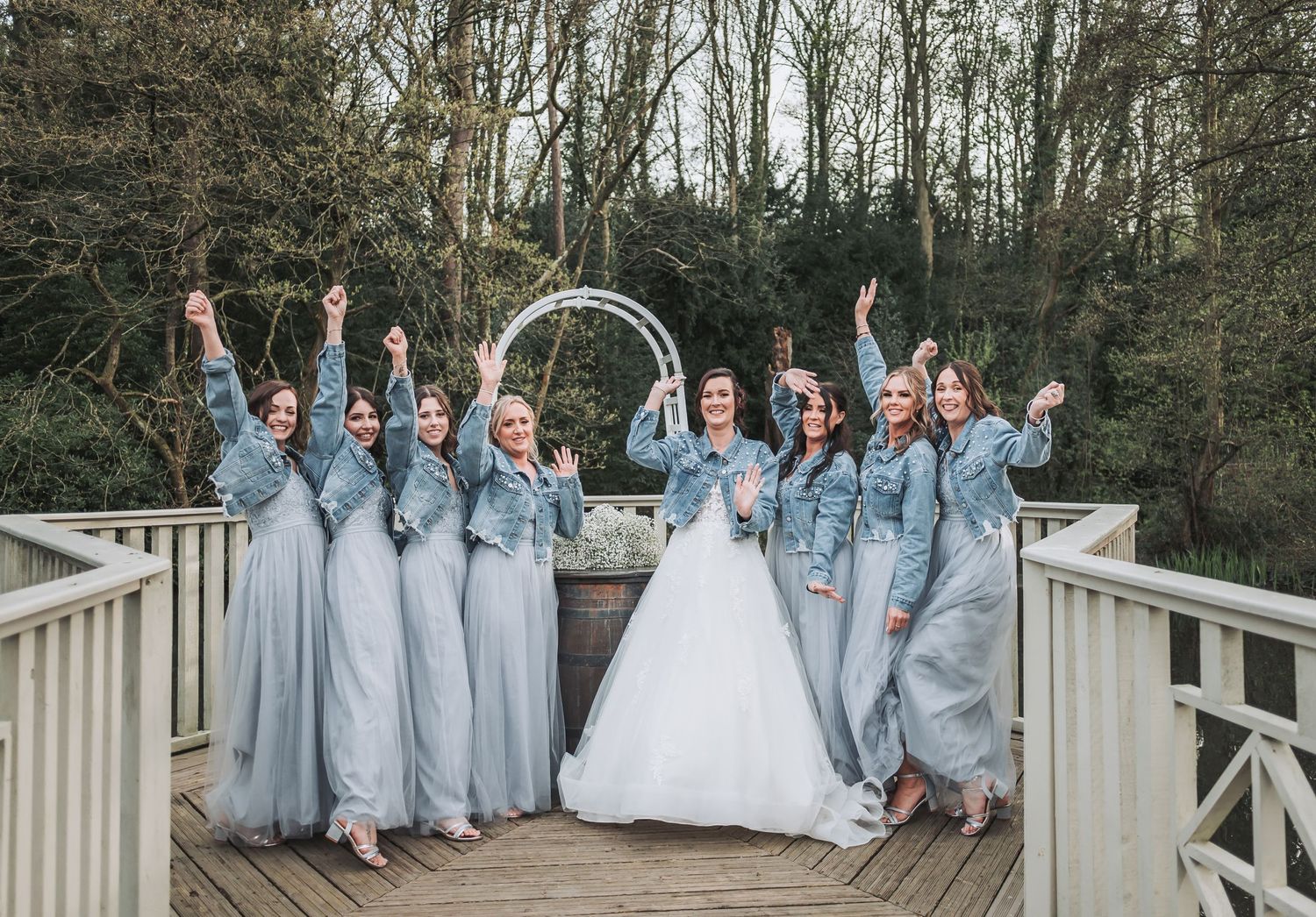 A bride in a denim jacket holds a bouquet while guests gather outside a historic stone building.