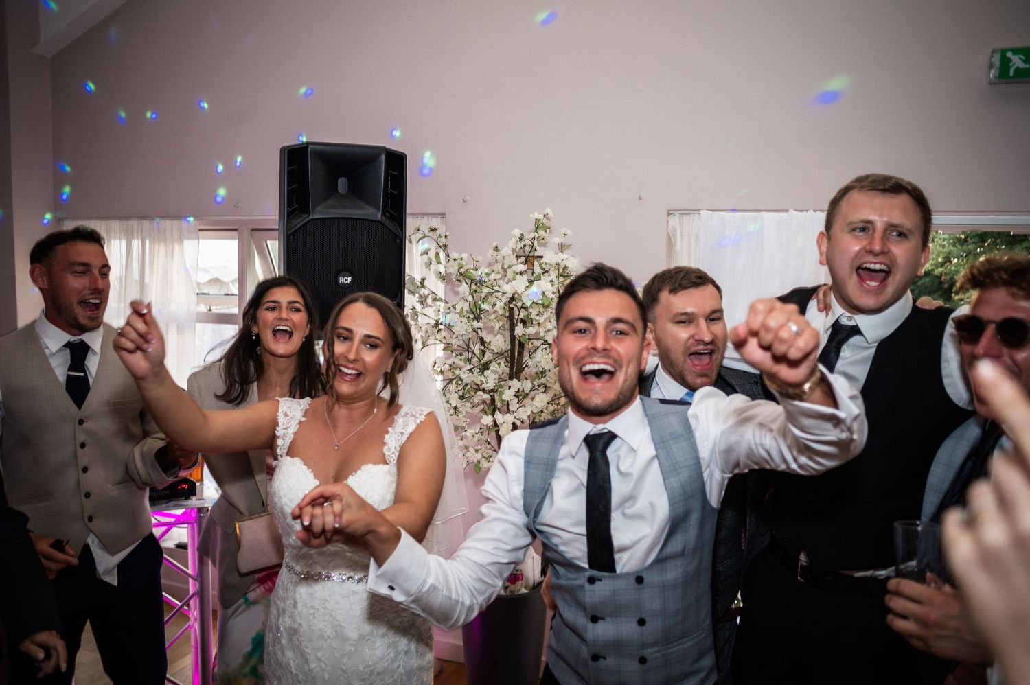 Wedding guests and party members celebrating enthusiastically on a dance floor with blue lighting.