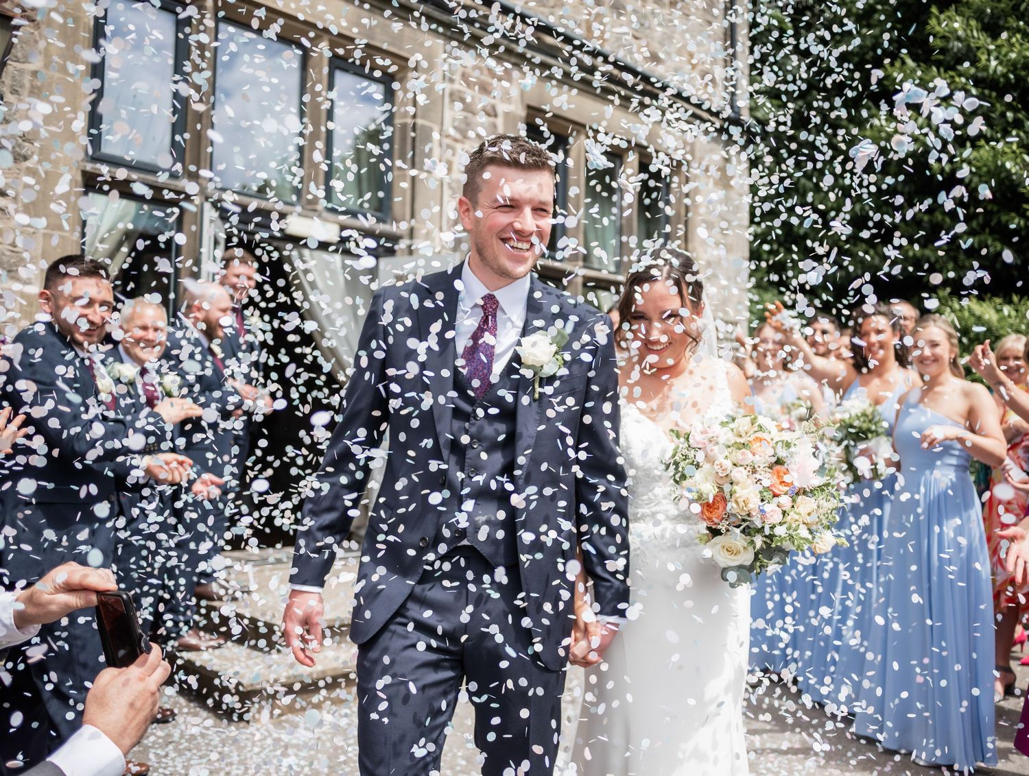Newlyweds celebrating under shower of white confetti outside stone building.
