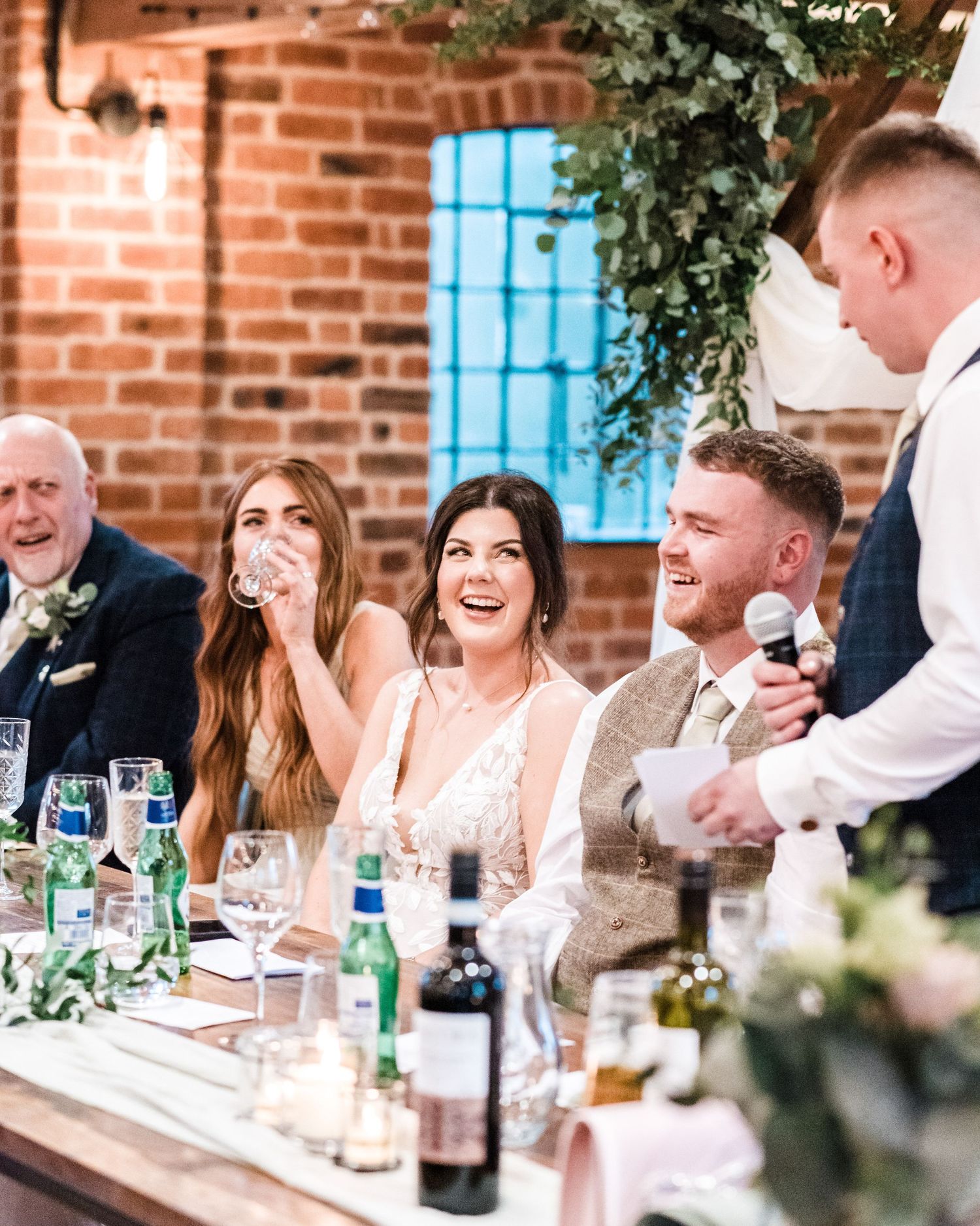 Wedding party laughing at head table during reception speeches in industrial brick venue.