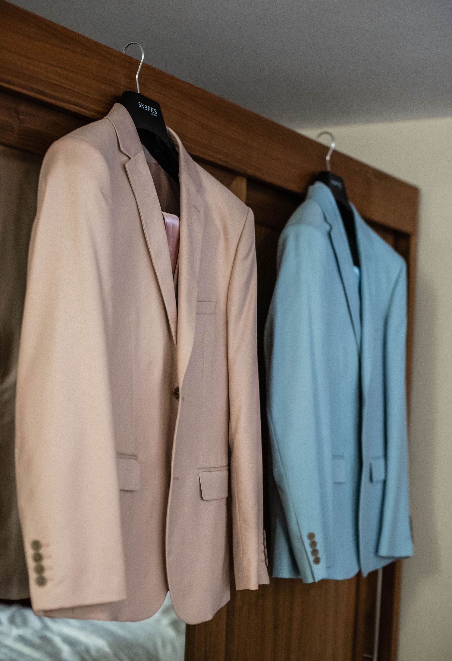 Two formal suit jackets in beige and blue hanging on wooden hangers.