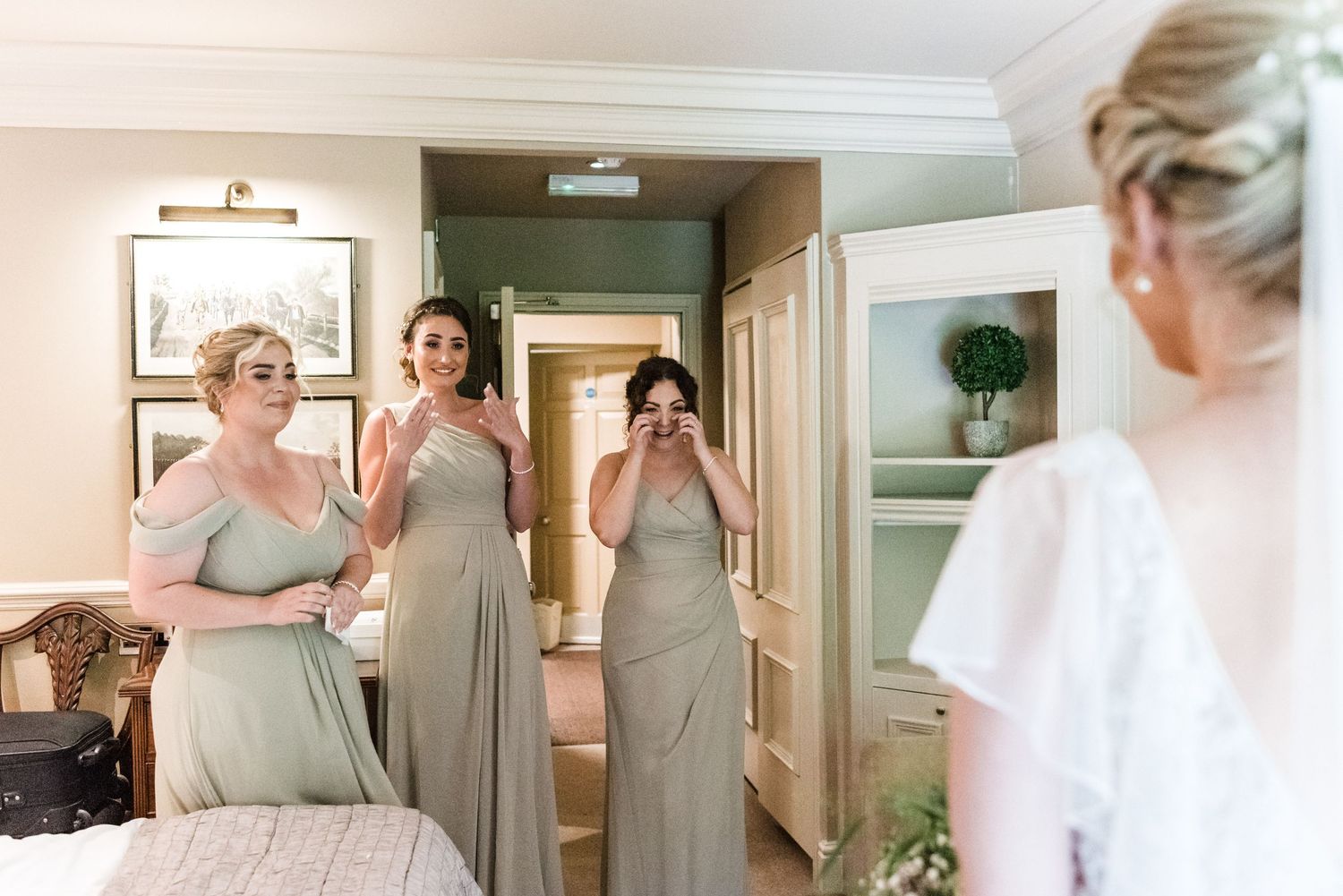 Bridesmaids in soft gray dresses share an emotional moment in a hotel room.