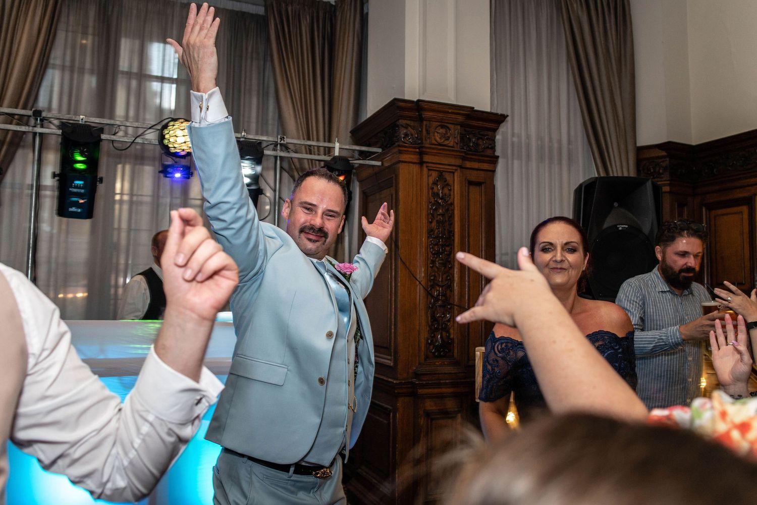 Wedding guests dancing and celebrating with raised hands in an elegant wood-paneled venue.