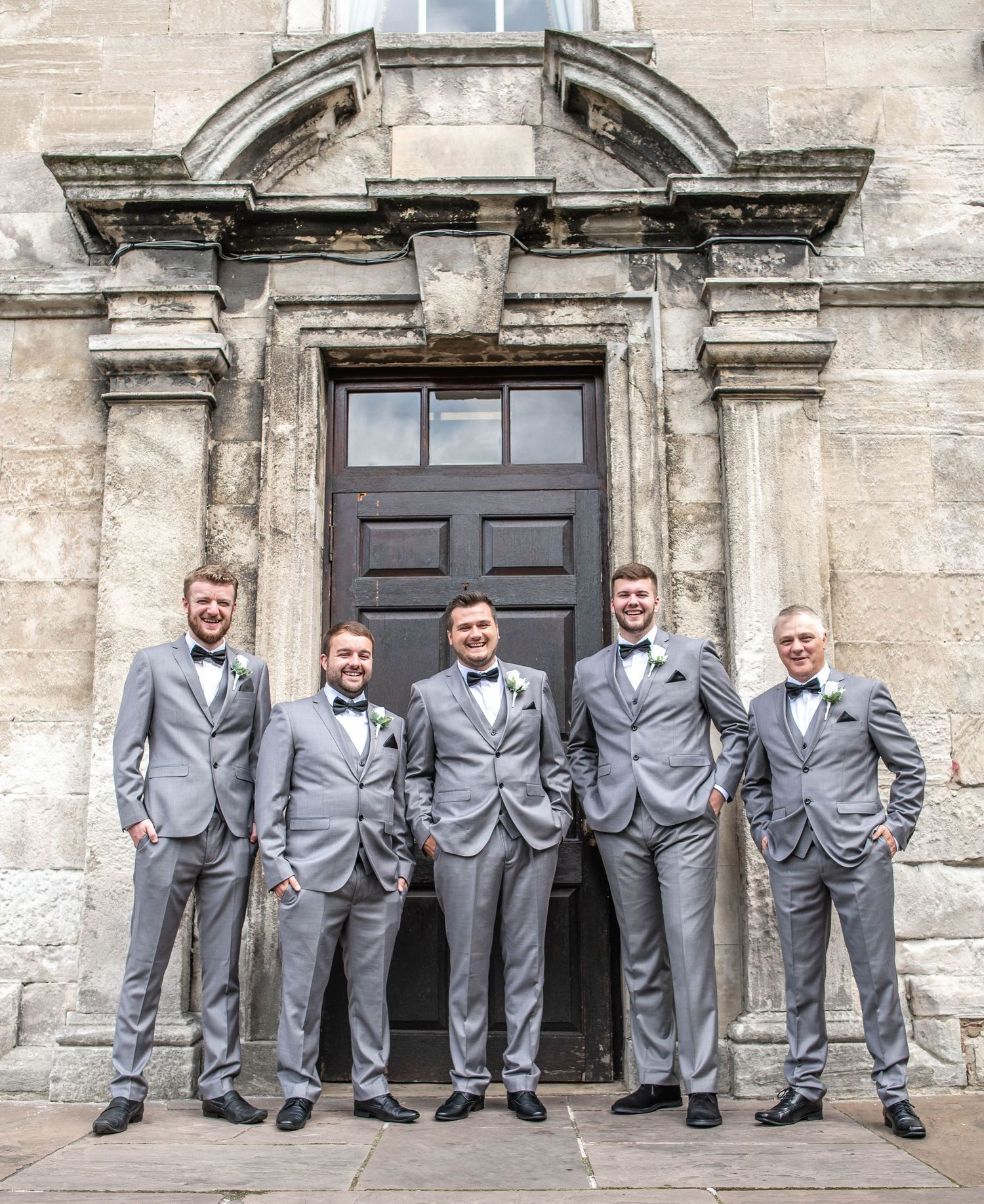 Groomsmen in matching grey suits posed in front of historic building entrance.