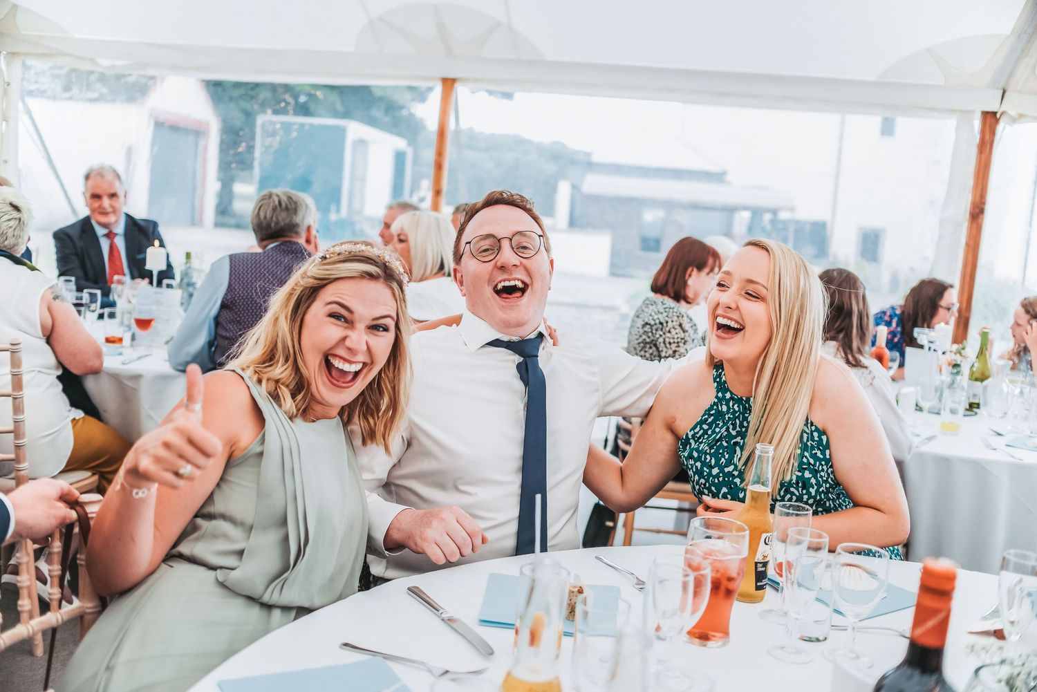 Wedding guests laughing and celebrating at reception table under white tent.