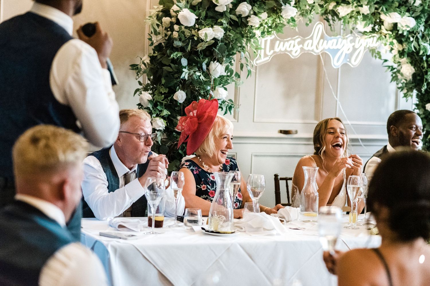 Reception guests laughing at dining table under floral arch with neon sign.