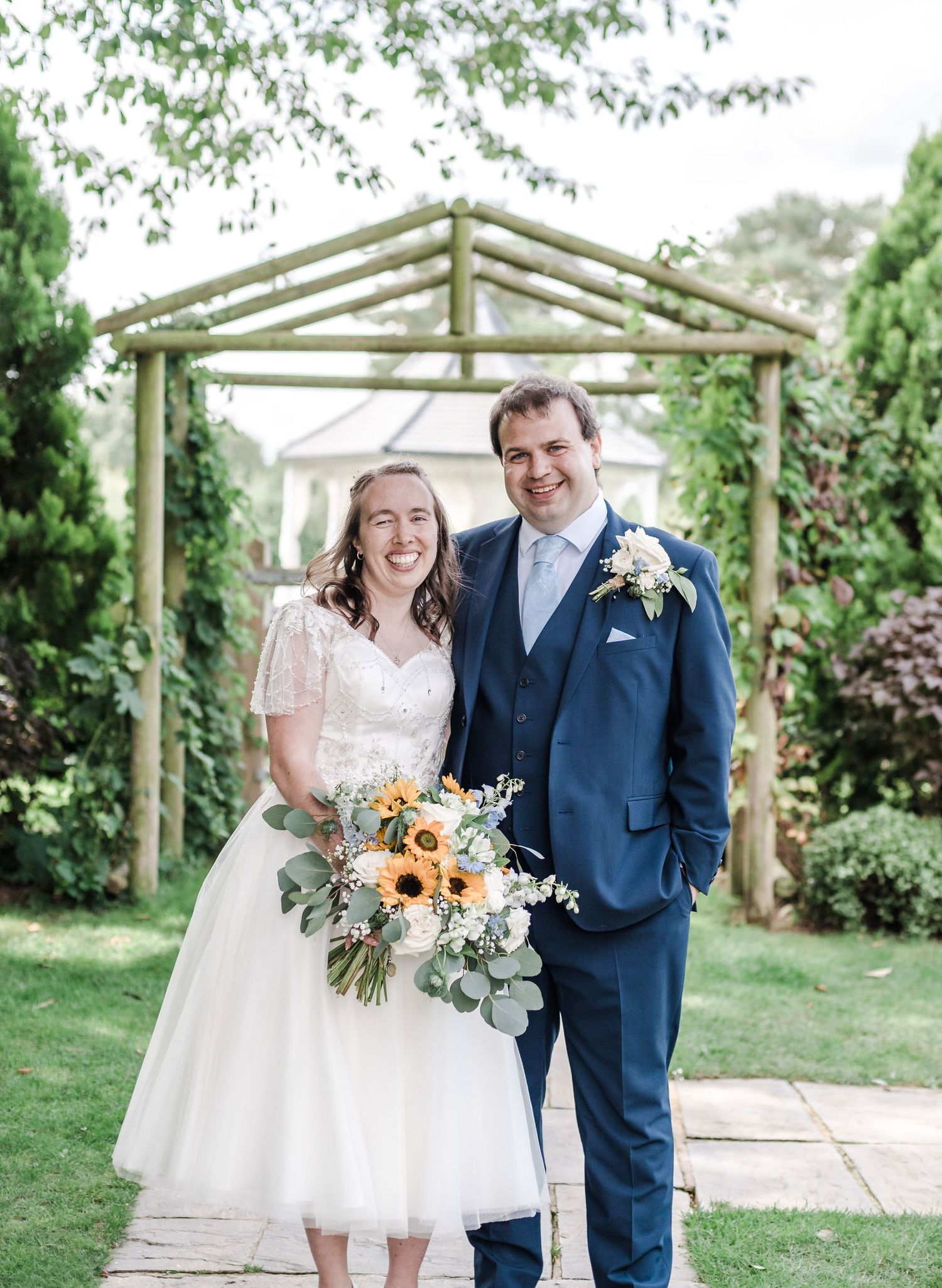 Wedding couple poses under wooden garden arbor with sunflower bouquet.