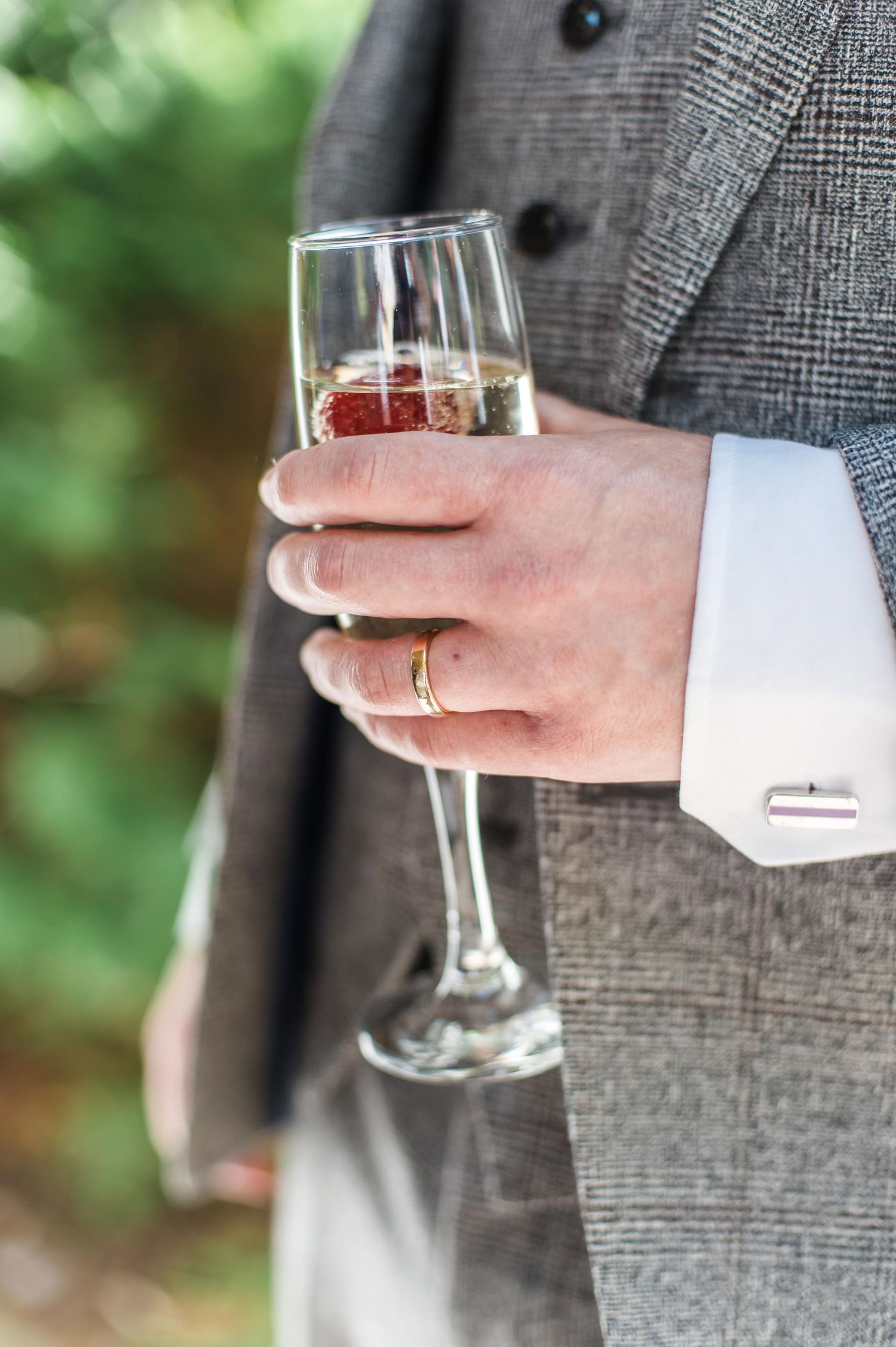 Close-up of hands holding champagne glass showing wedding ring and grey suit sleeve.