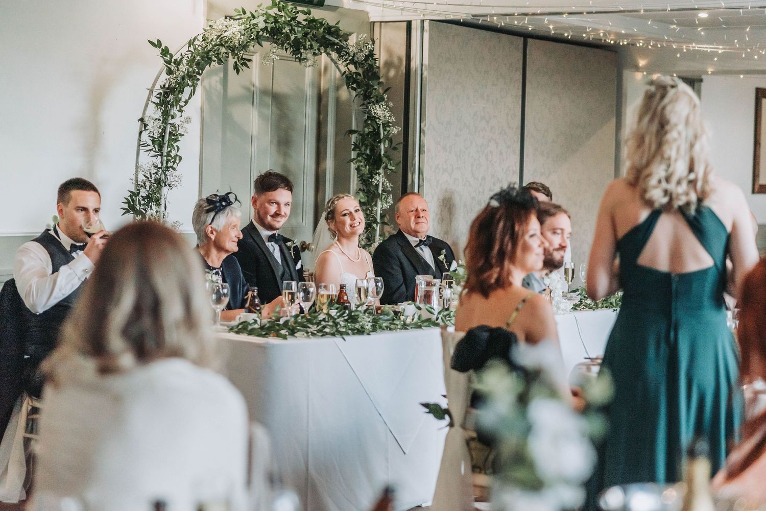 Wedding reception dinner with guests seated at tables decorated with greenery and string lights.