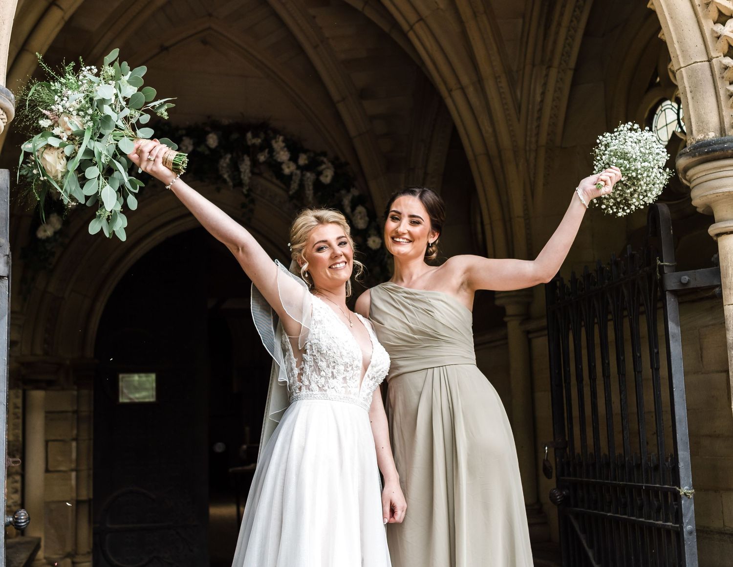 Two wedding party members celebrating with raised bouquets under stone archway.