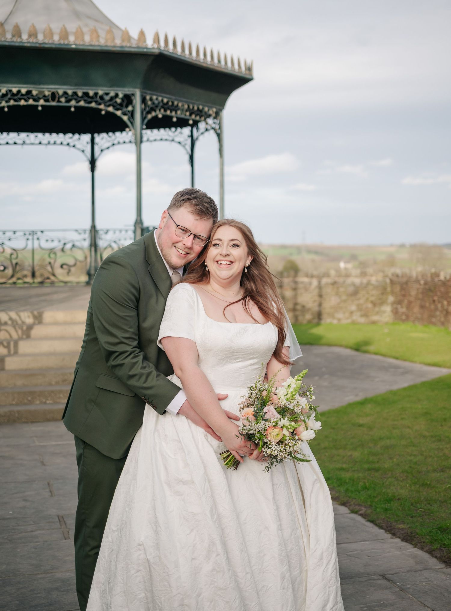 A wedding couple embraces under a Victorian-style gazebo on a grassy hilltop overlooking a scenic landscape.