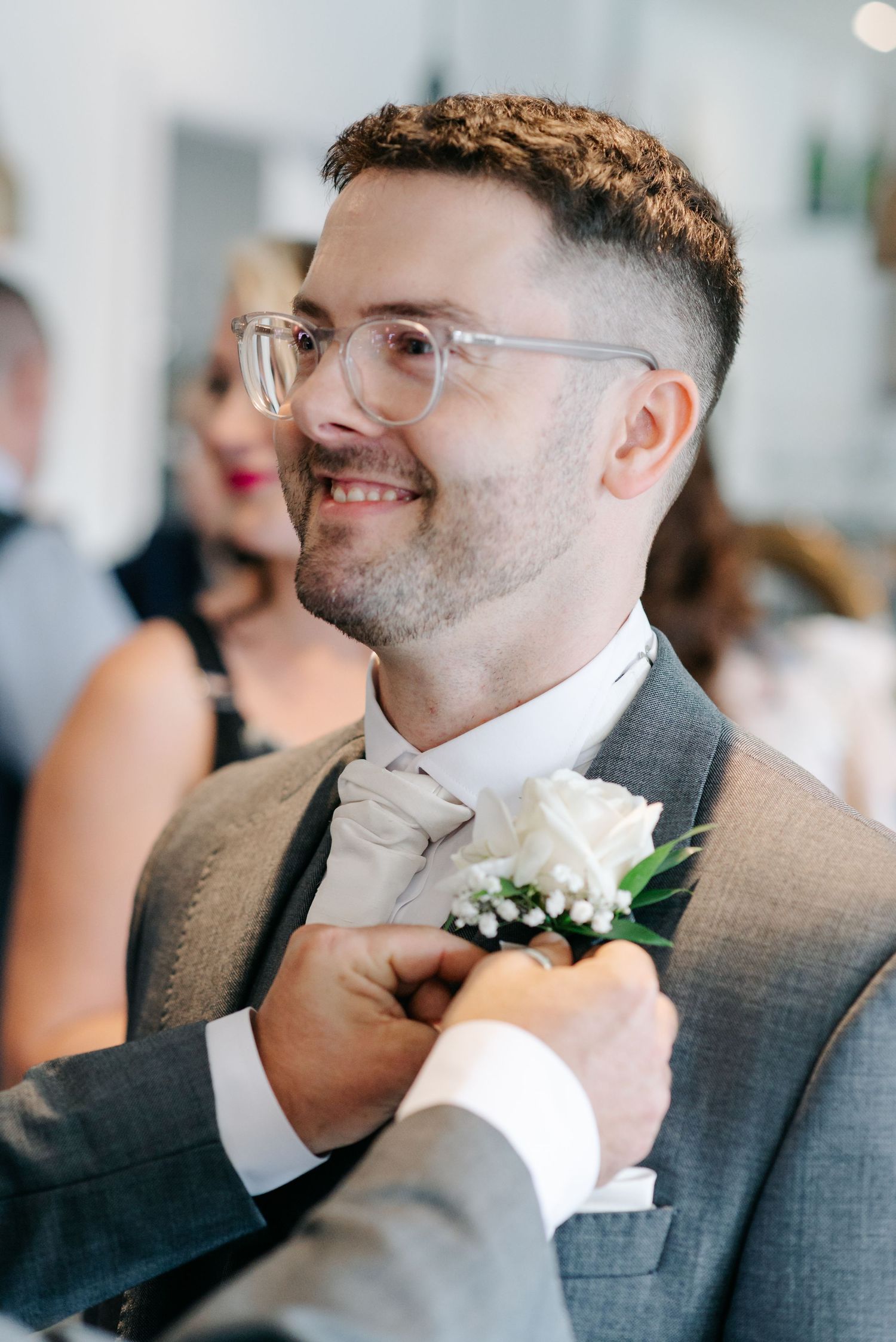A person wearing glasses and a grey suit adjusts their white boutonniere flower while smiling.