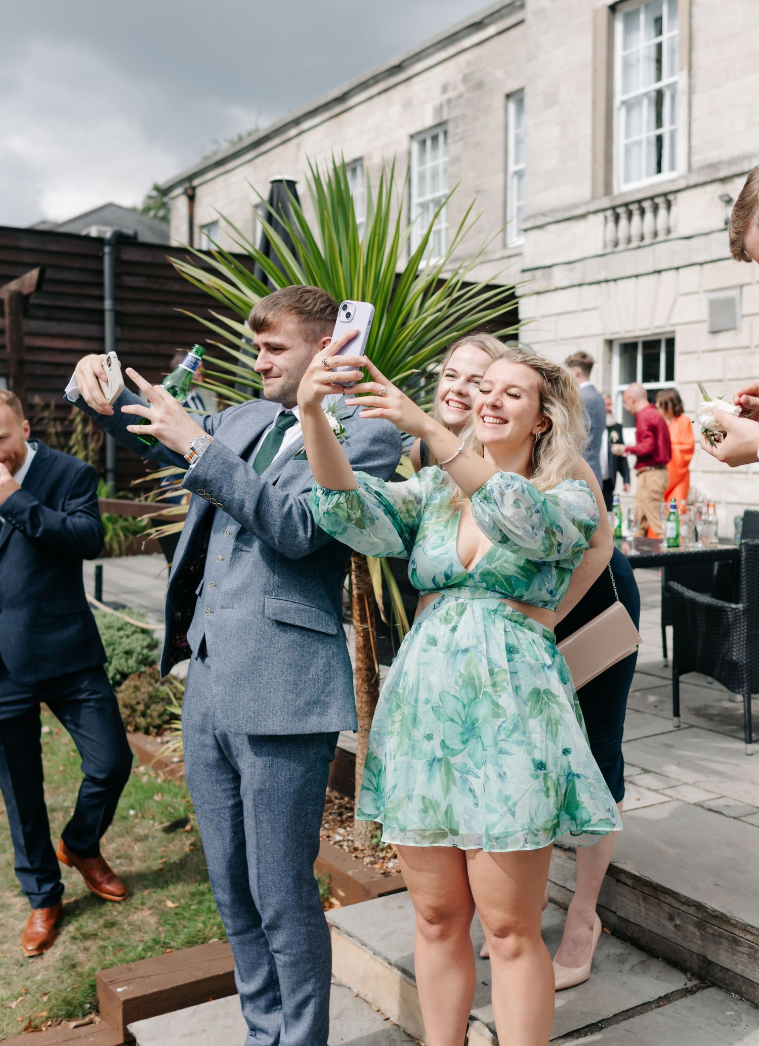 Wedding guests in formal attire celebrate and take selfies outside a stone building.
