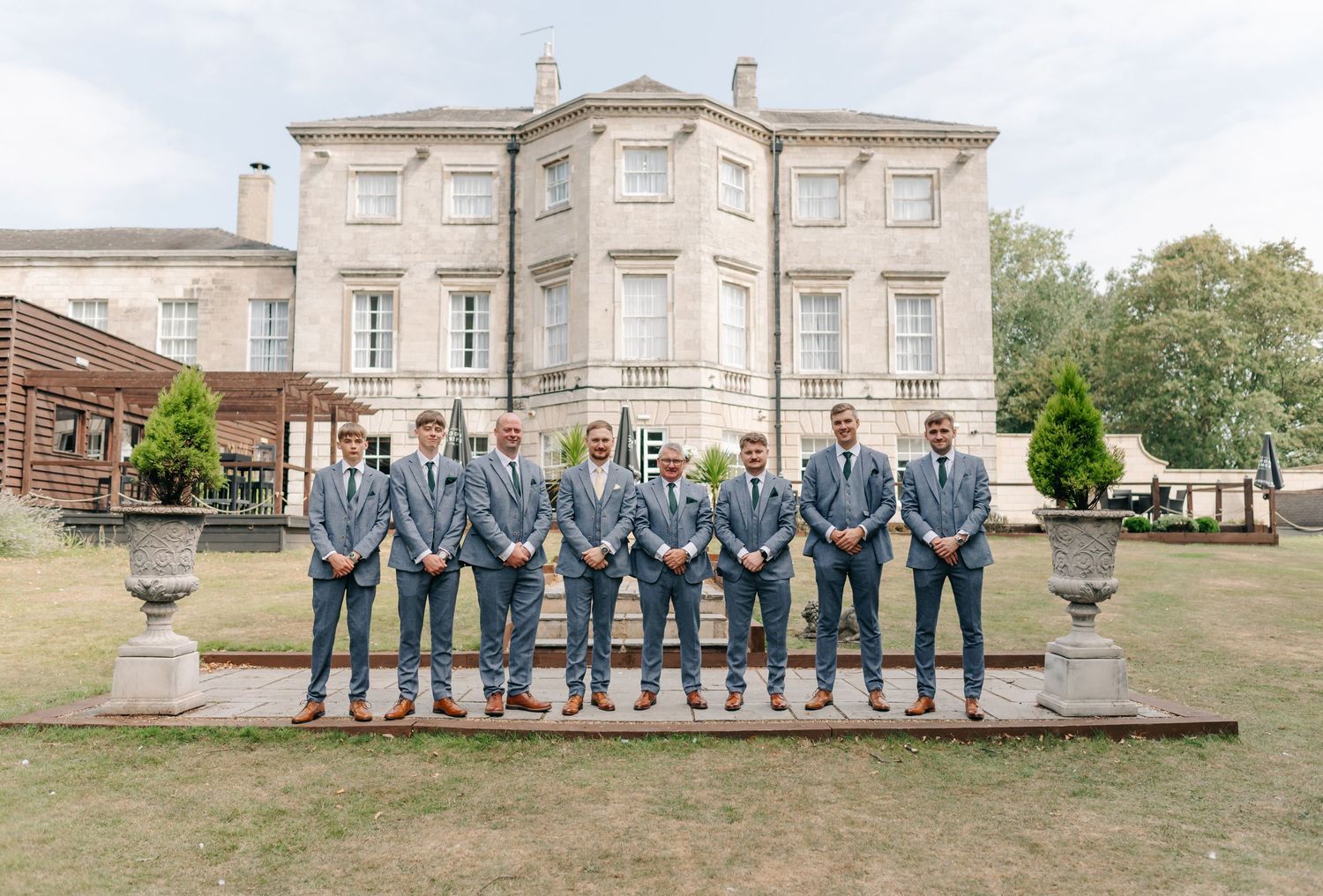 A group of groomsmen in matching gray suits stand in a line in front of a grand historic mansion.